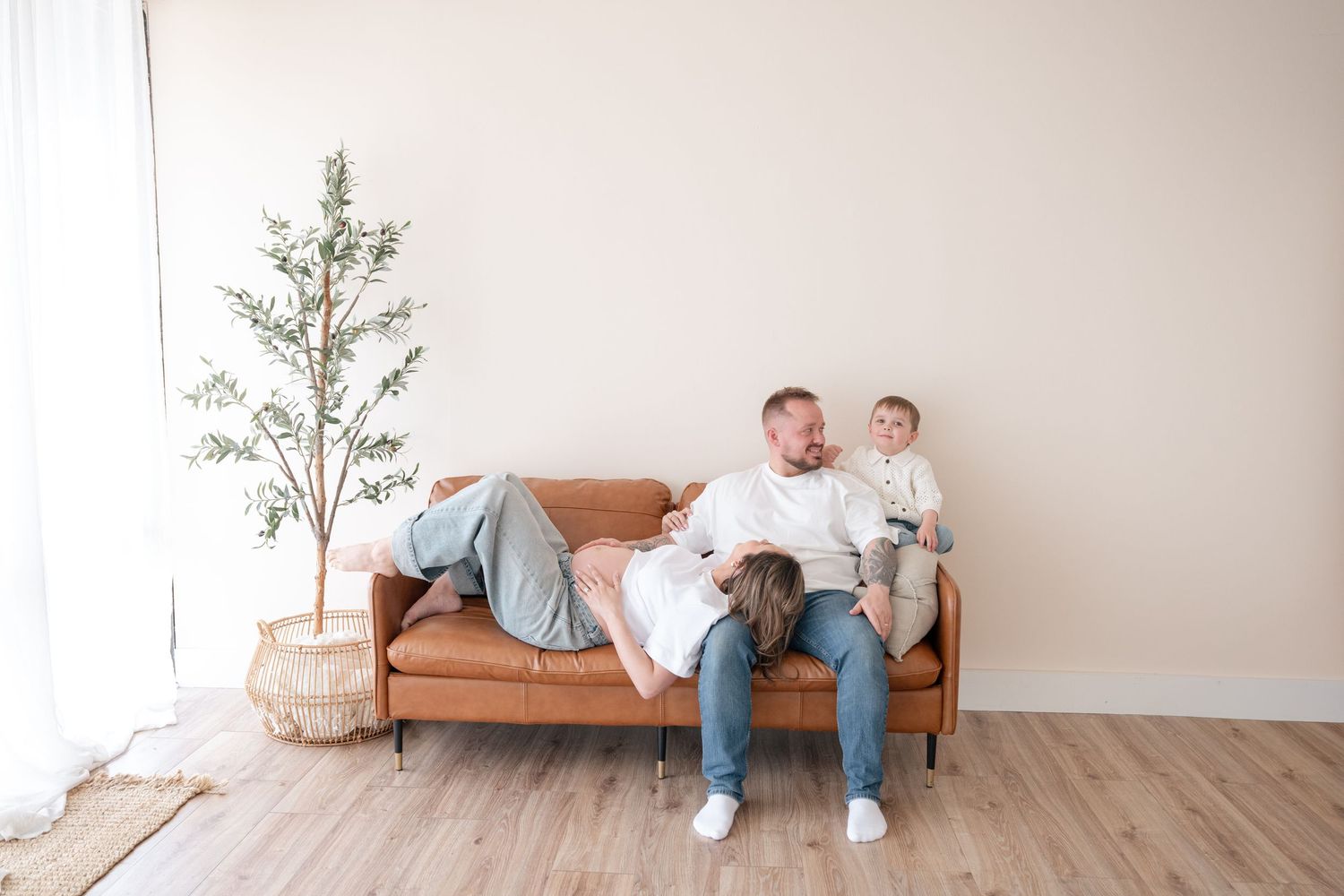 Family relaxing together on a brown leather sofa in a bright modern living room with hardwood floors and a potted tree.