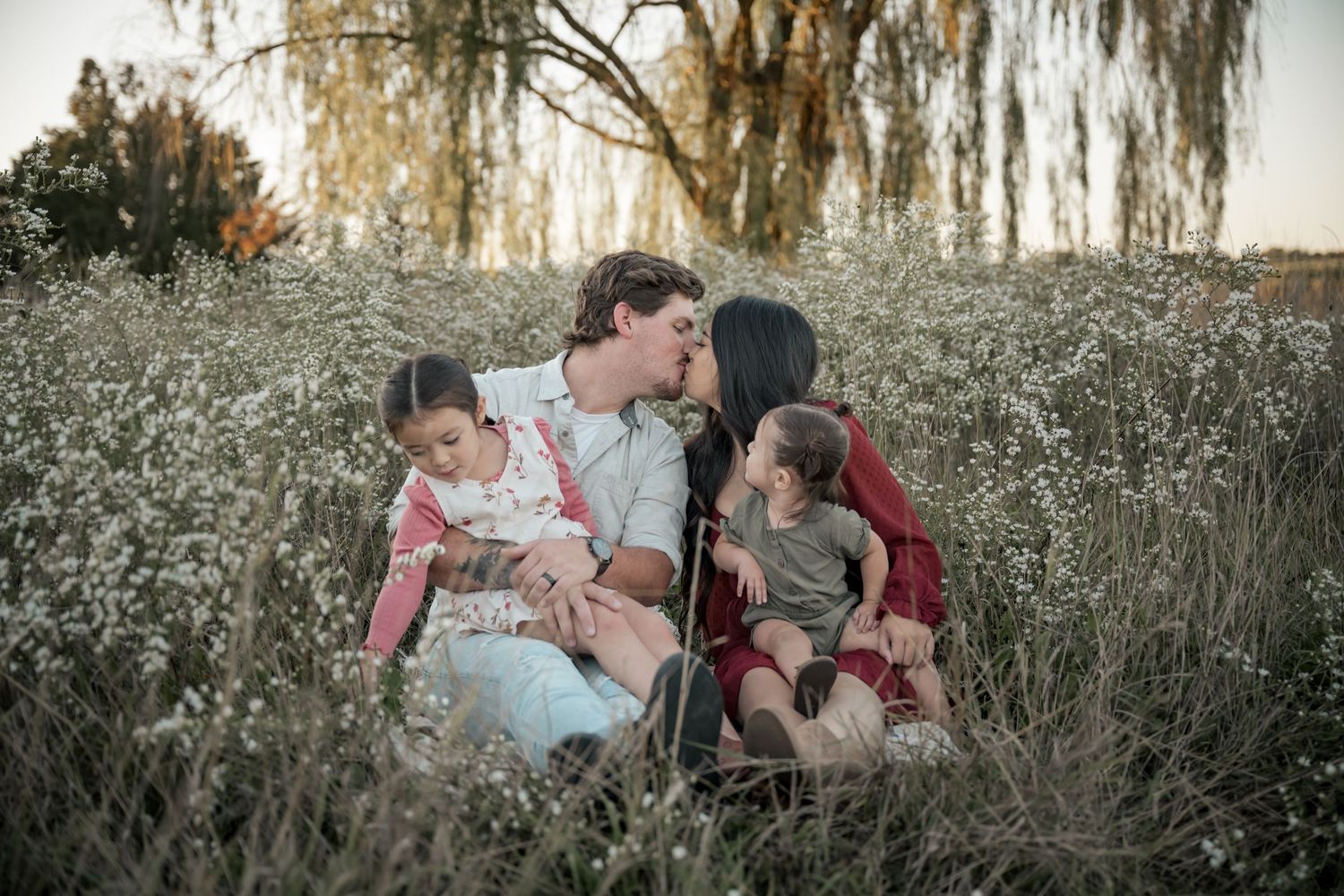 Family of four sitting in a wildflower field, parents kissing while holding their two young children at golden hour.