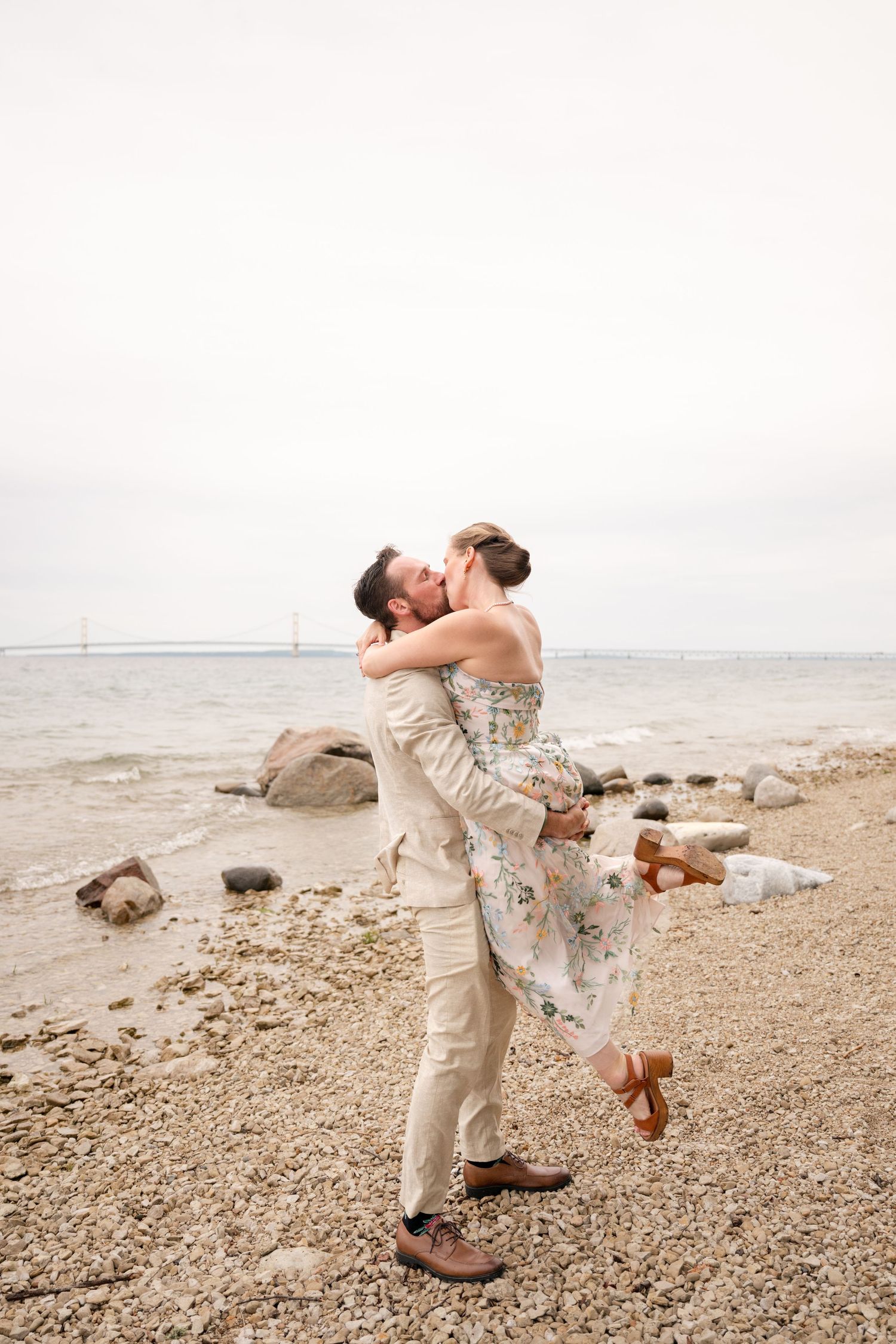 A couple shares a romantic kiss as the man lifts his partner on a rocky shoreline with a bridge in the distance.