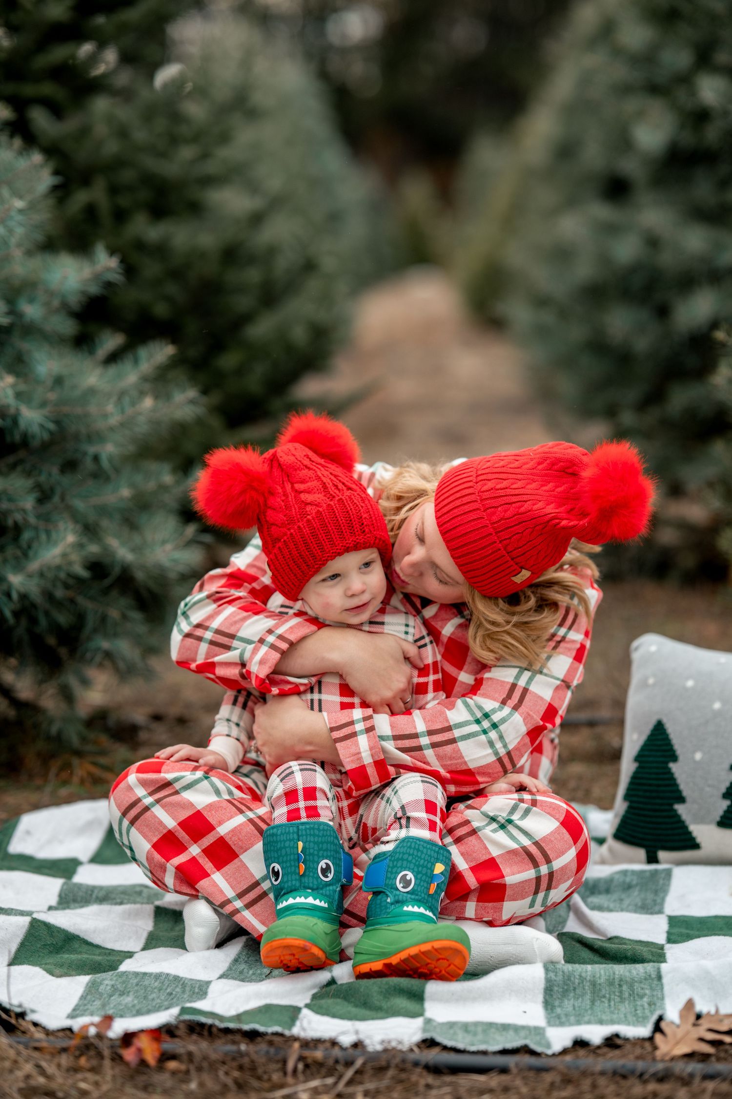 Mother and child in matching red plaid pajamas and red pom-pom hats hugging at a Christmas tree farm.