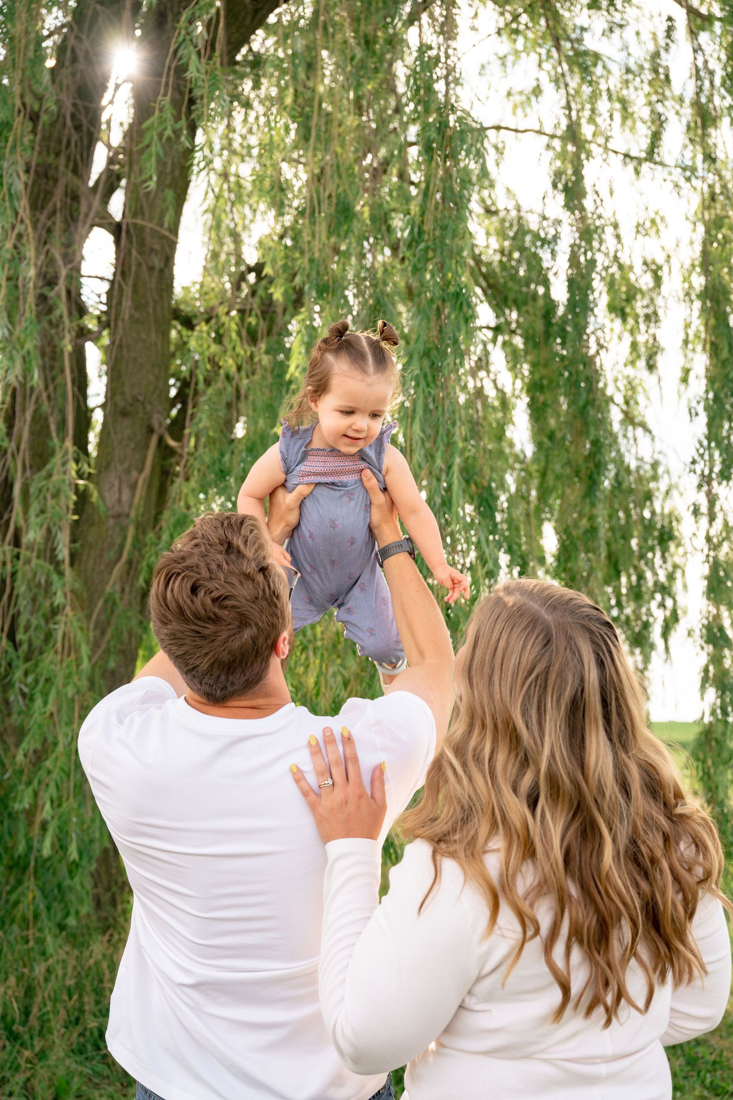 Parents lifting their happy toddler in the air under a willow tree outdoors.
