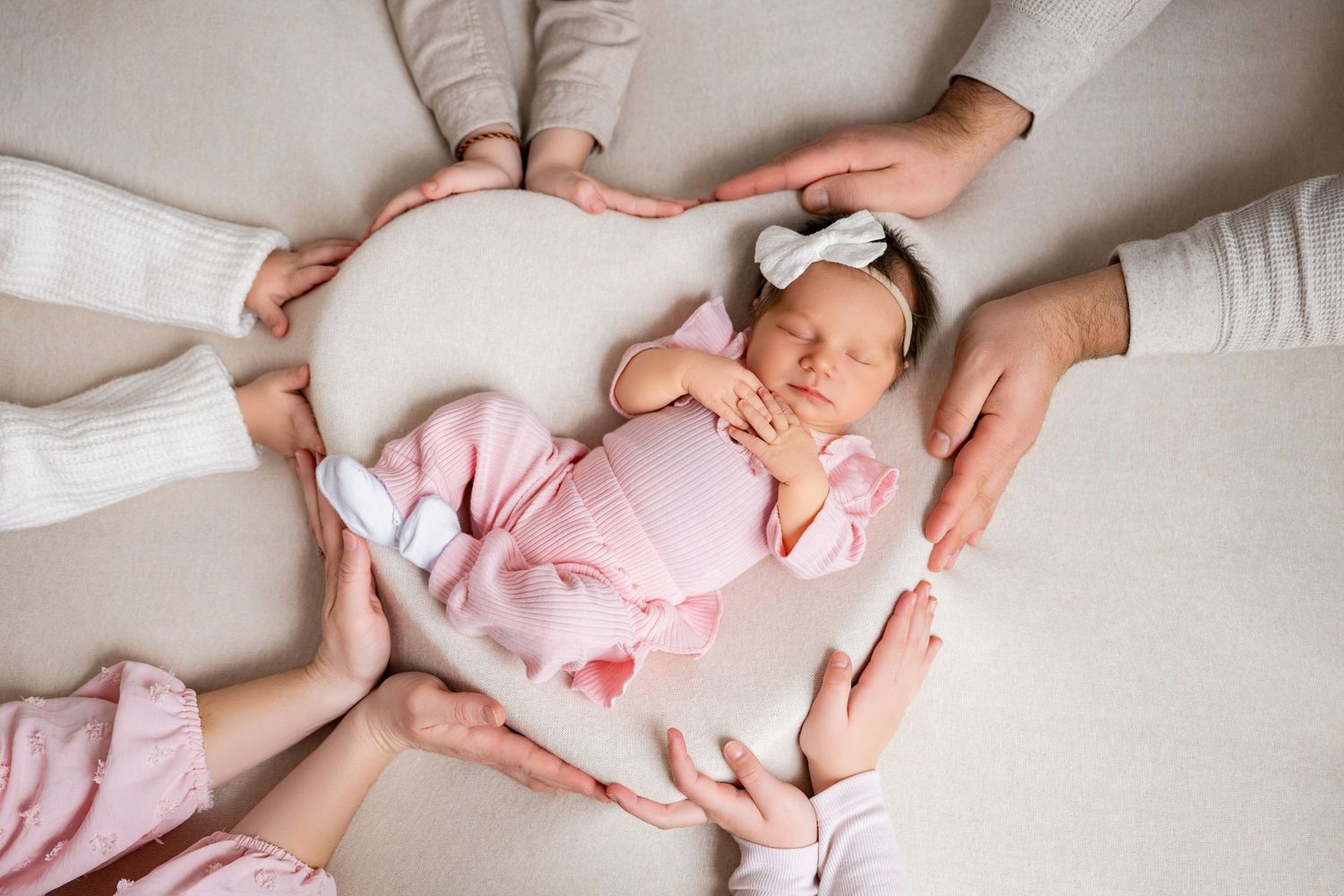 Newborn baby girl in pink outfit sleeping peacefully, cradled by multiple family hands forming a heart shape.