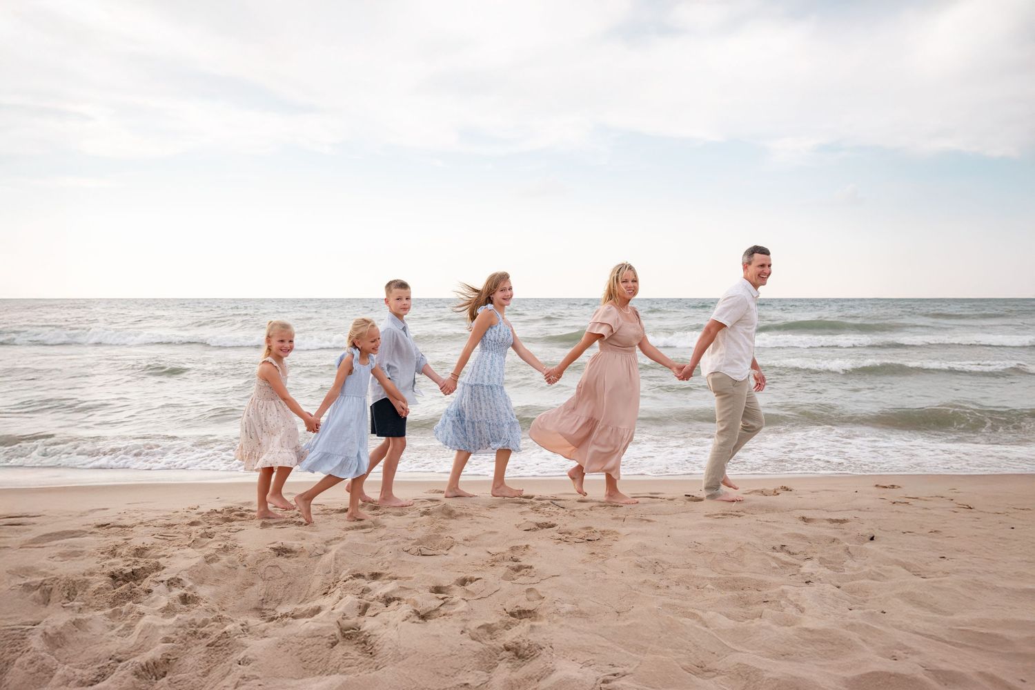 Happy family of six holding hands and running together along a sandy beach at sunset.