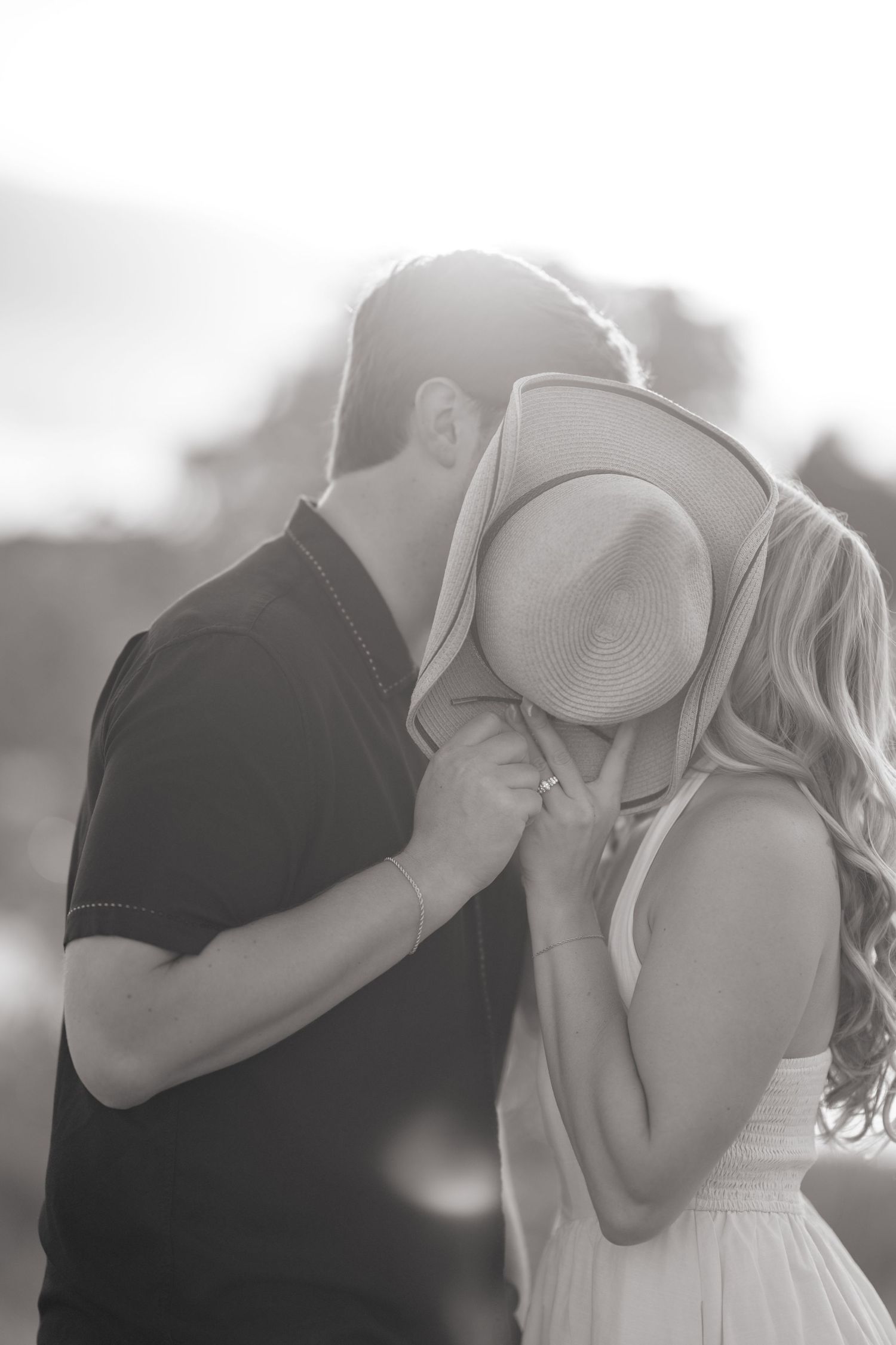 Black and white photo of a couple sharing a romantic kiss hidden behind a straw hat in soft backlit sunlight.