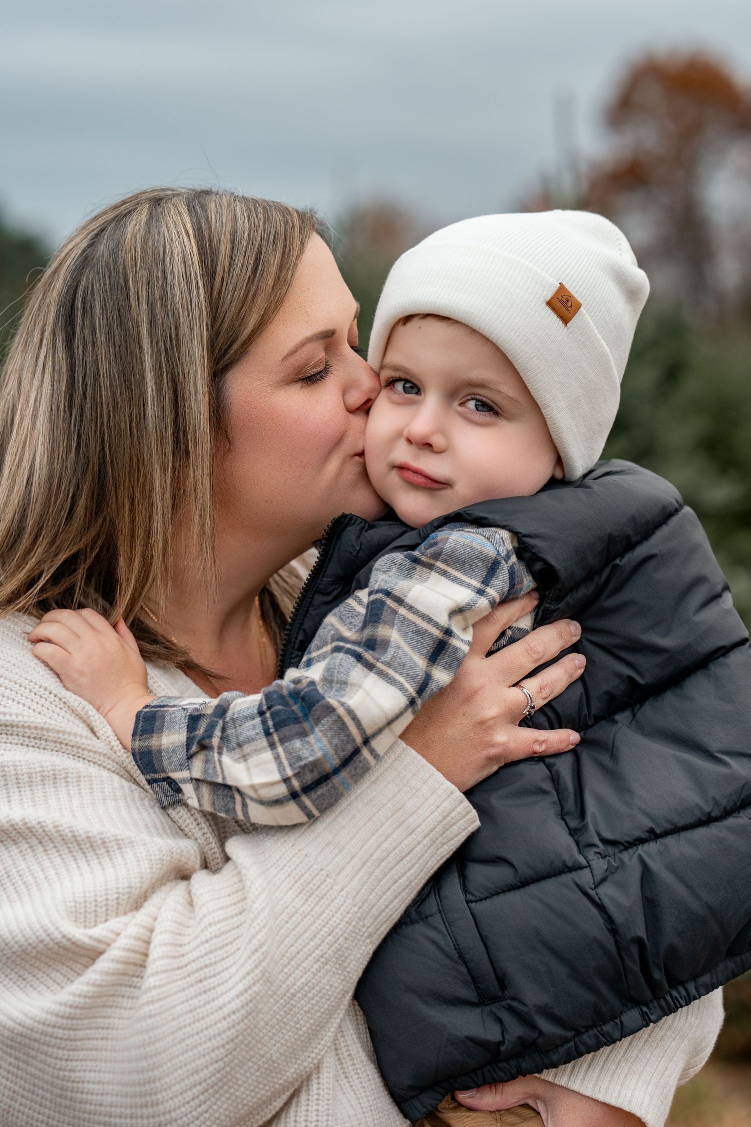 A mother kisses her young child on the cheek outdoors in autumn, child wearing a white beanie and black puffer jacket.