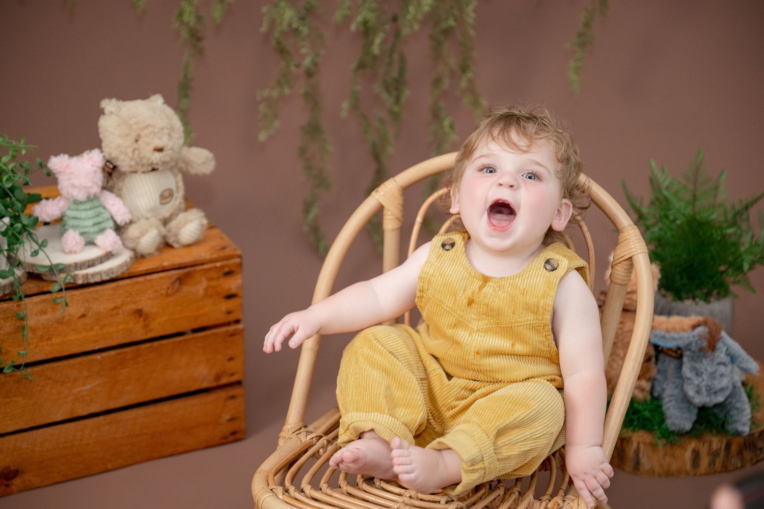 Happy toddler in yellow romper sitting in wicker chair with stuffed animals and plants in background.