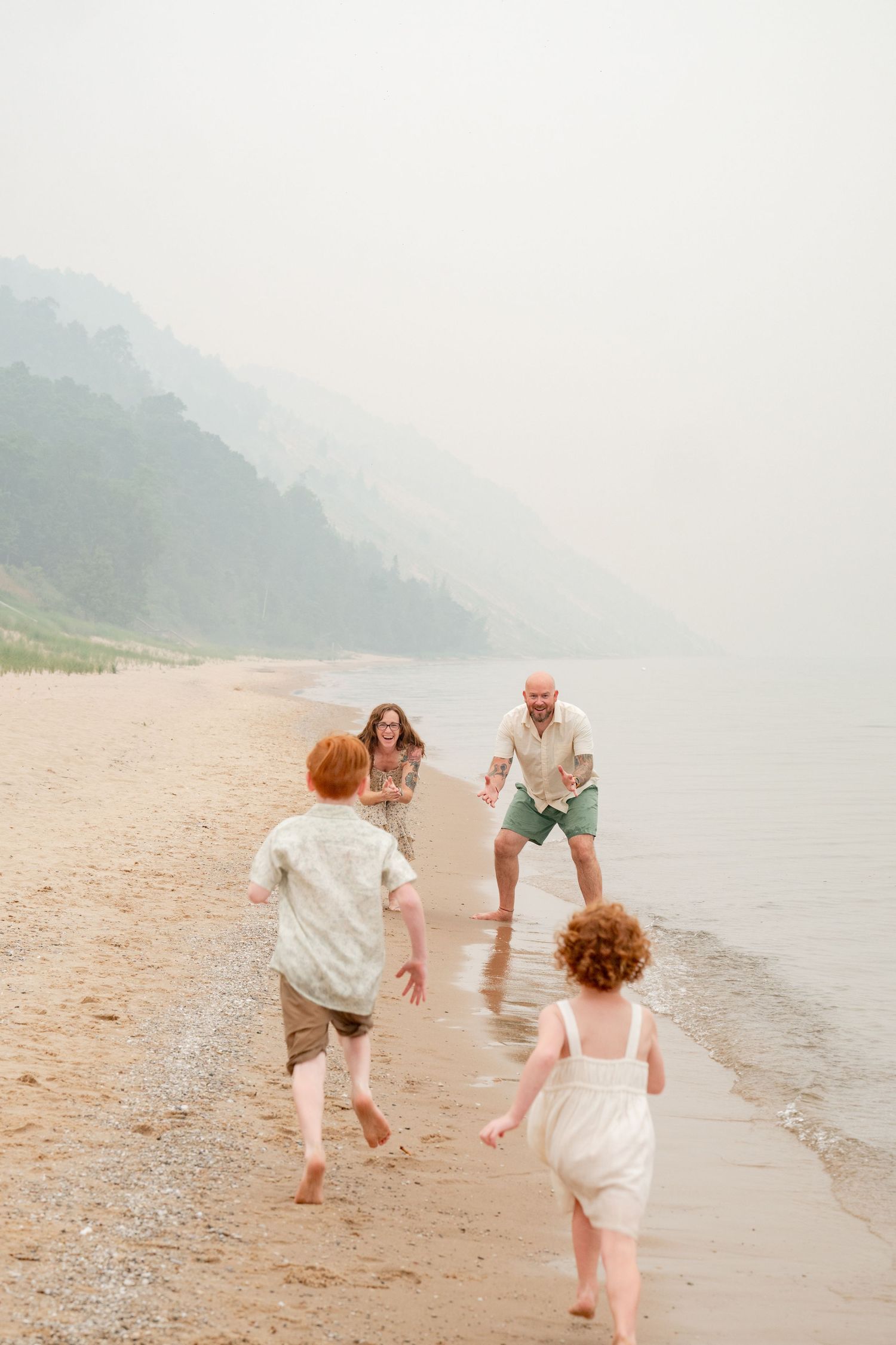 Two red-haired children run toward their parents along a misty, foggy shoreline beach during family playtime.