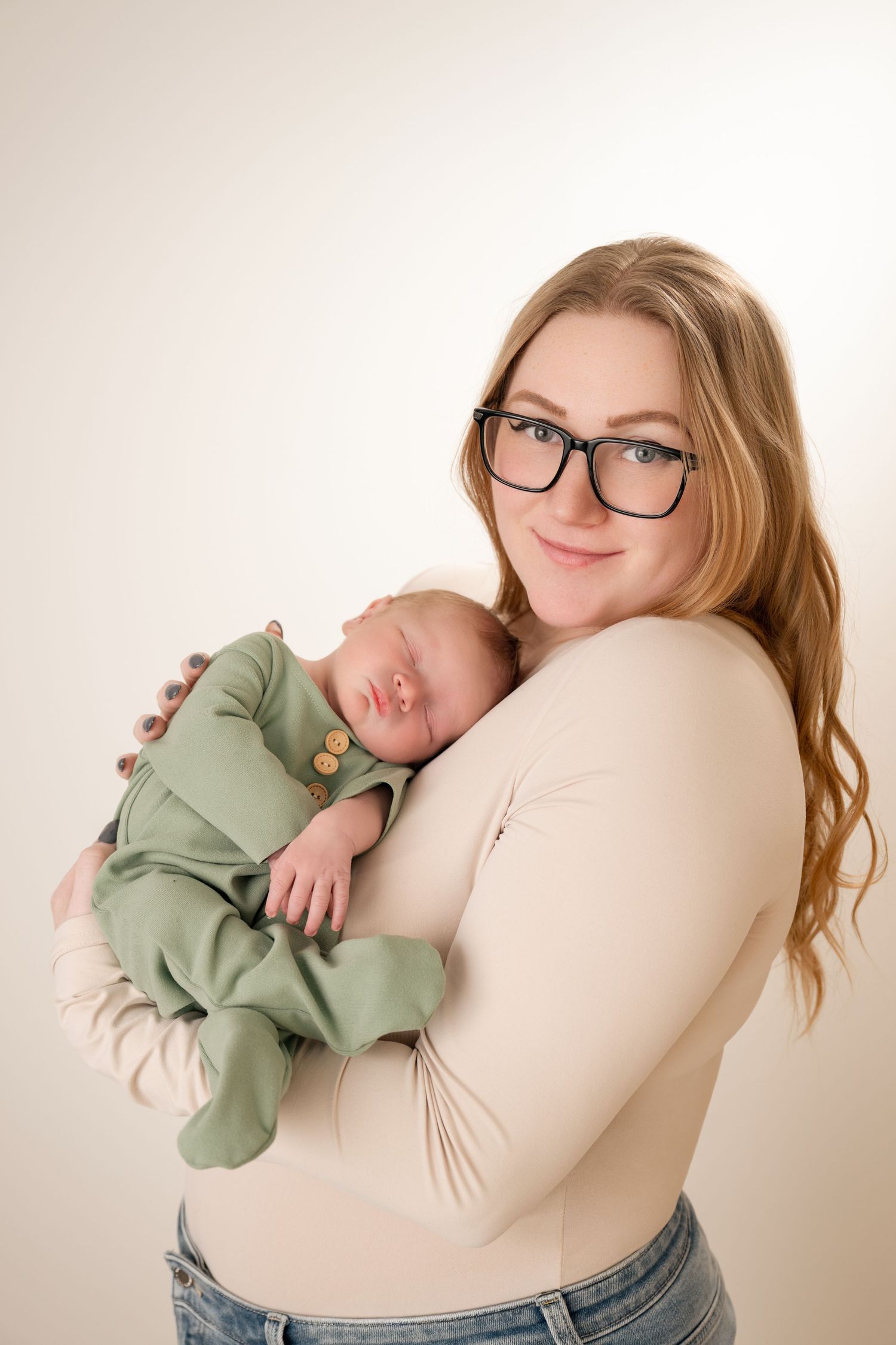 A smiling mother with glasses holds her sleeping newborn baby dressed in a green outfit against a light background.