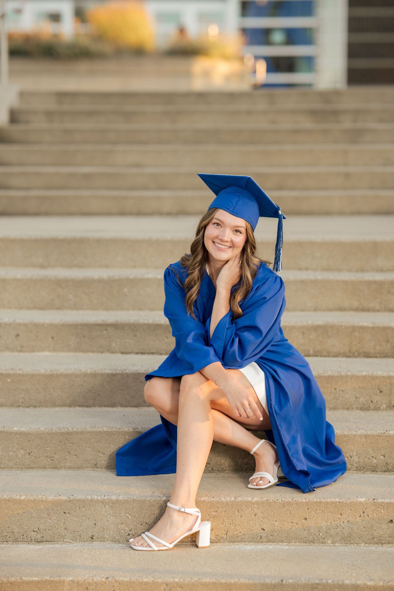 Smiling young woman in blue graduation cap and gown sitting on outdoor steps wearing white heeled sandals.