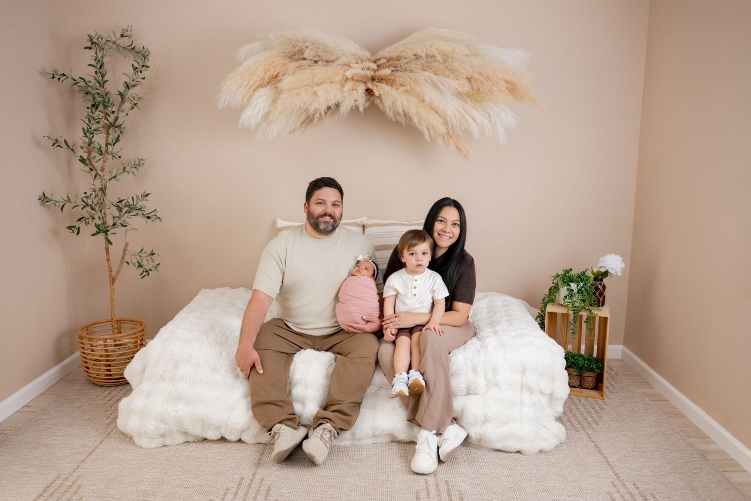 Family of three posing on a white fluffy bed in a boho-styled studio with pampas grass decor and neutral tones.