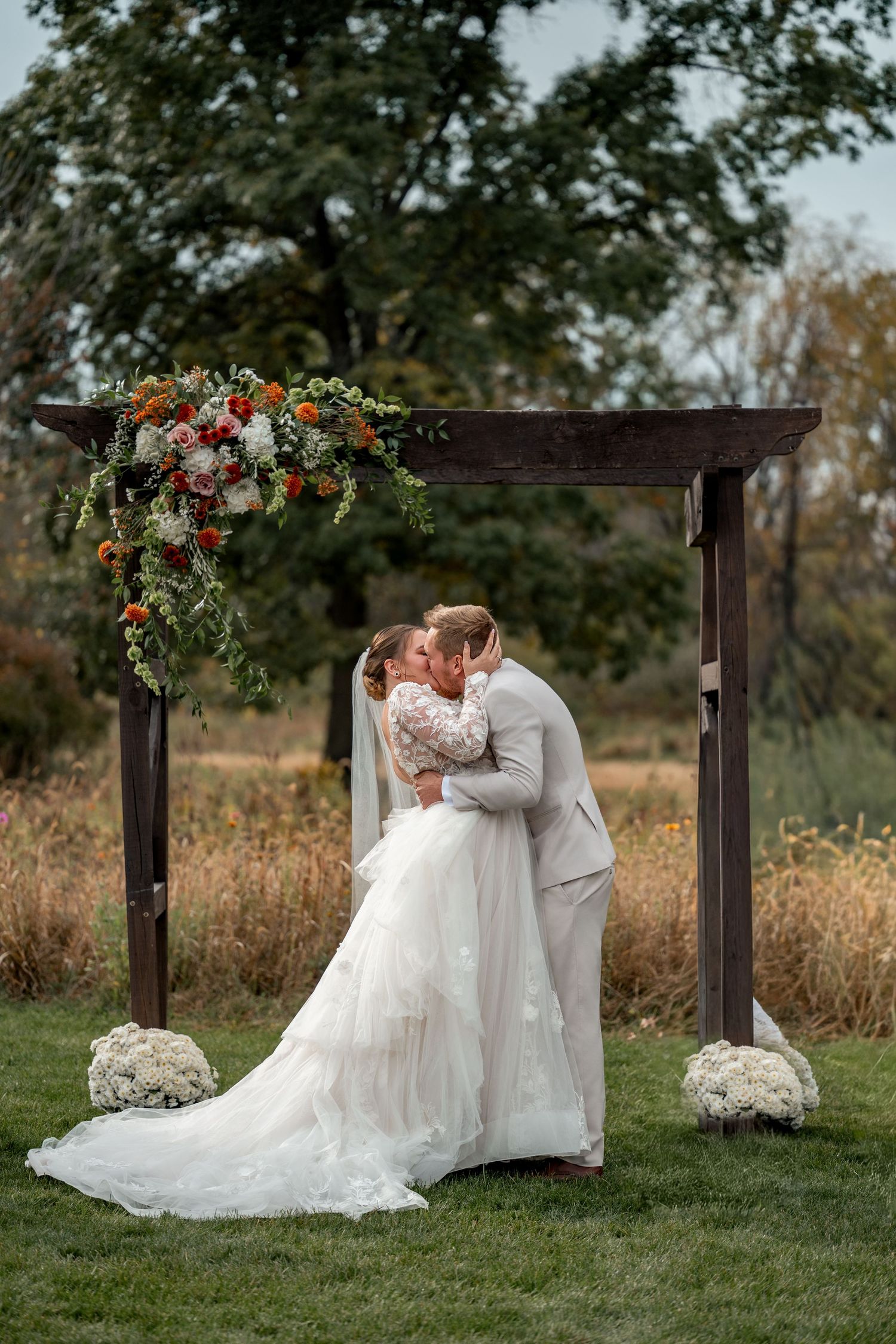 Bride and groom share first kiss under rustic wooden arch adorned with red and white florals at outdoor fall wedding.