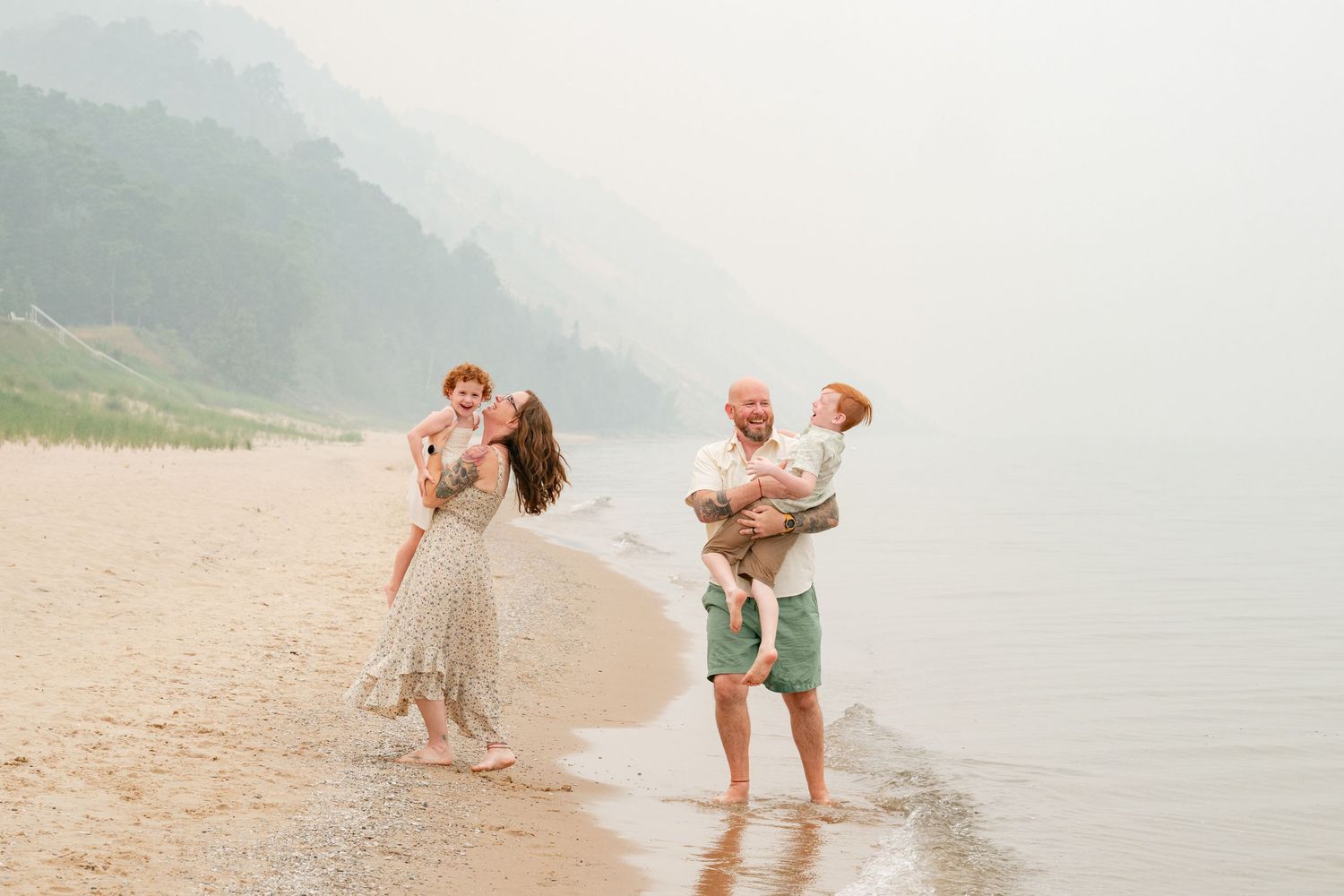 Happy family on a misty beach, mother and father each carrying a toddler while standing by the shoreline.