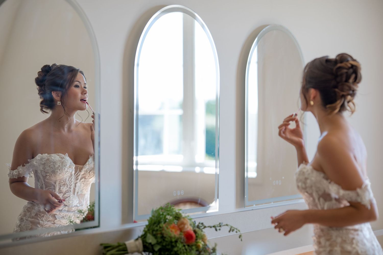 Bride in lace wedding dress applying perfume while admiring her reflection in arched mirrors.