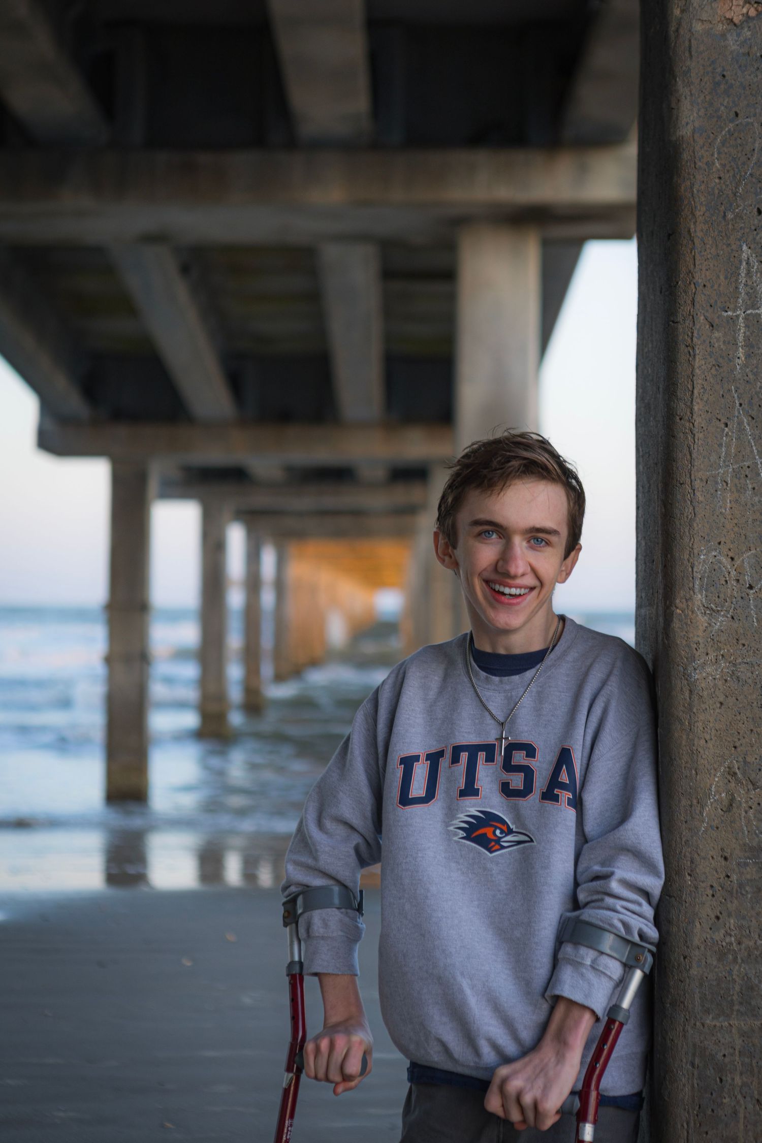 Smiling young man on crutches wearing a UTSA sweatshirt, leaning against a pillar under a pier.