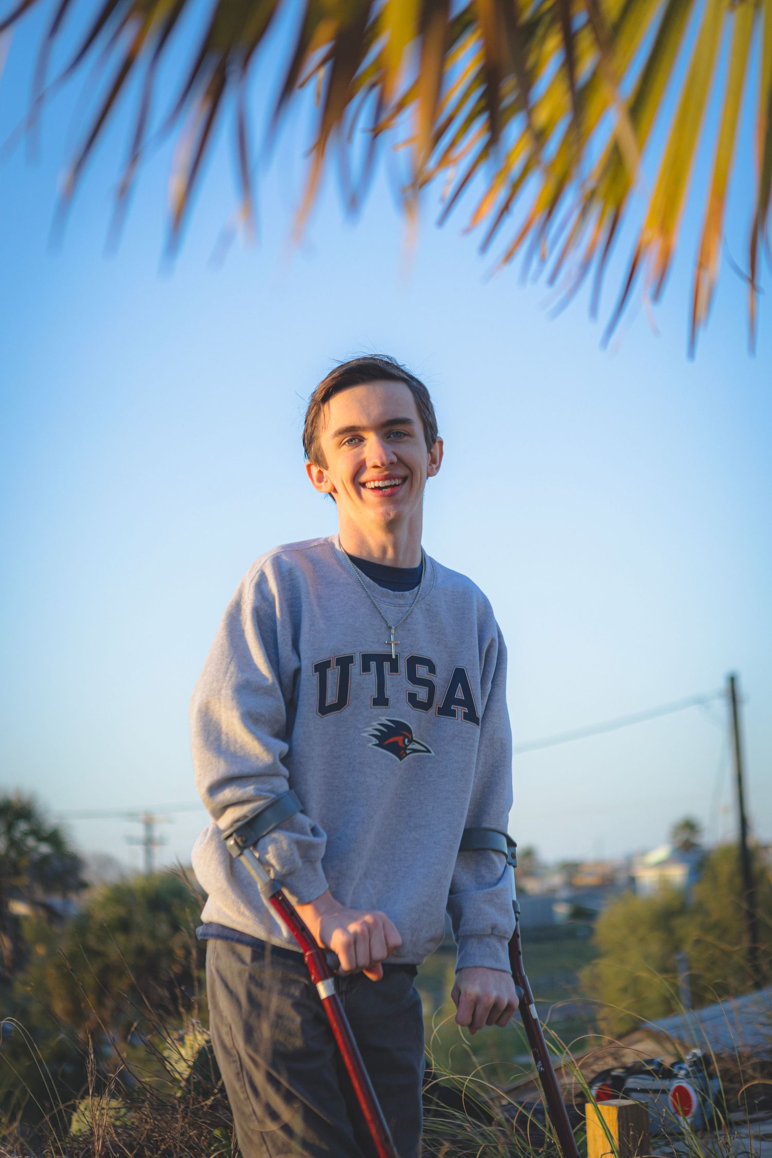 Young man smiling outdoors in UTSA sweatshirt, holding a golf club under palm fronds at golden hour.
