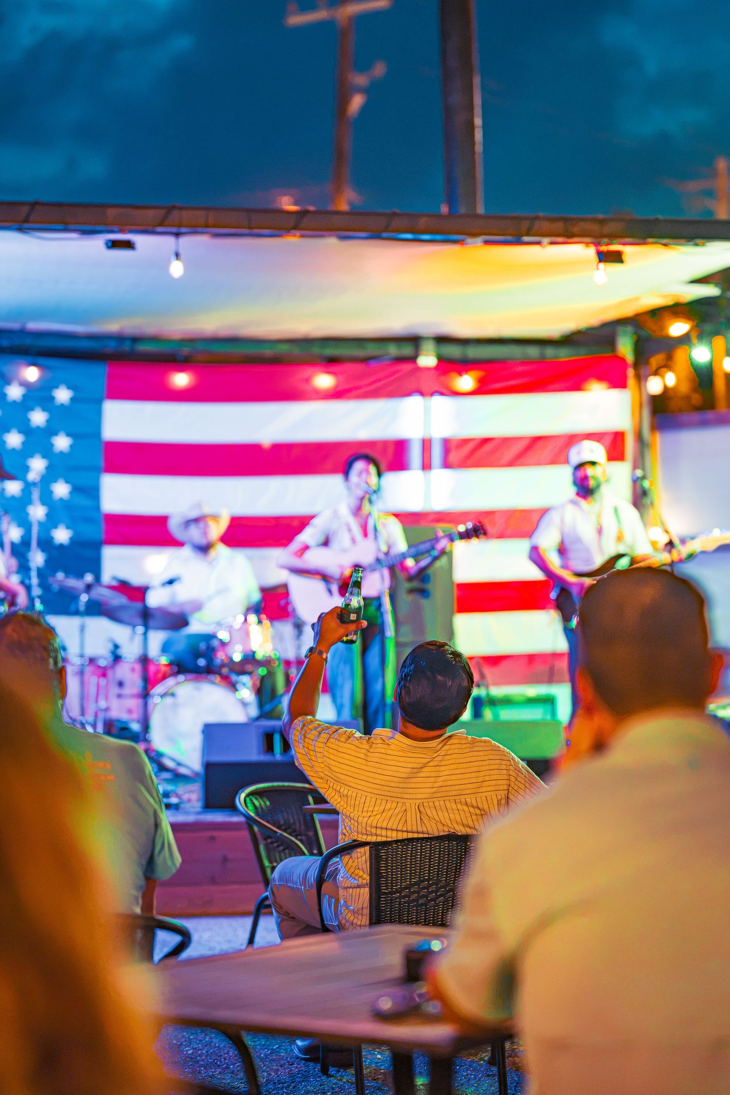 Live band performs on outdoor stage with American flag backdrop as audience watches under evening lights.