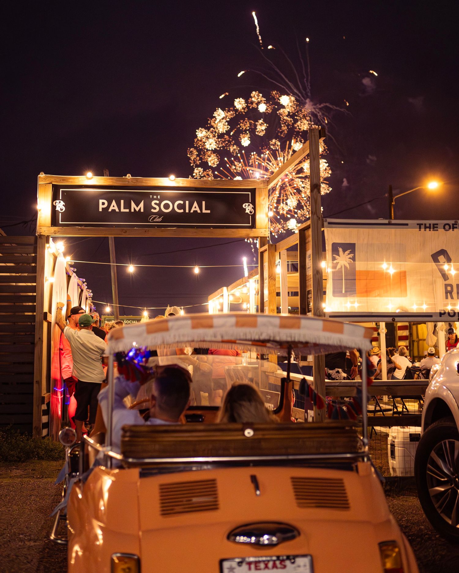 Vintage convertible car parked outside Palm Social Club at night with fireworks lighting the sky.