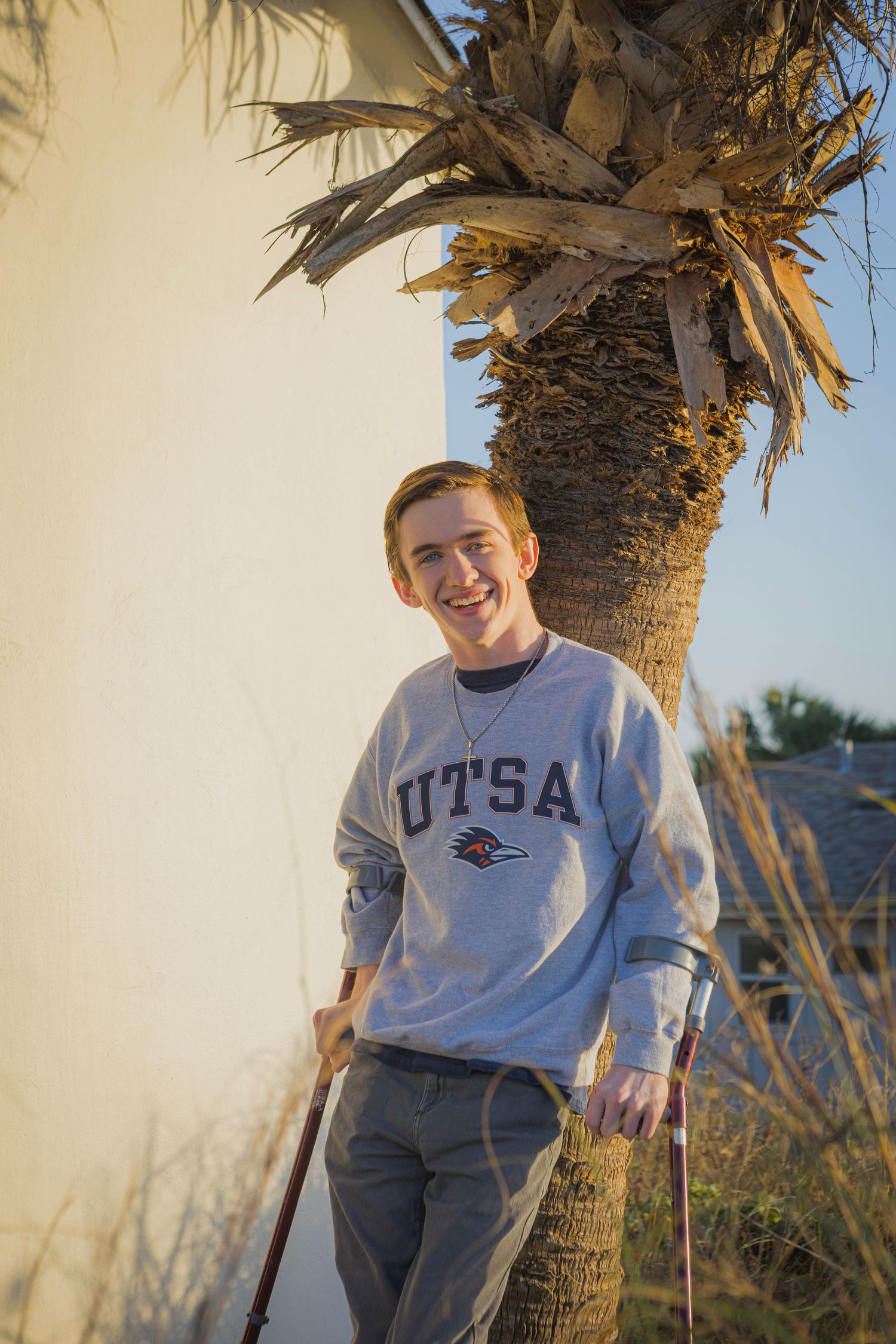 Young man smiling in UTSA sweatshirt leaning on crutches beside a palm tree in warm golden sunlight.