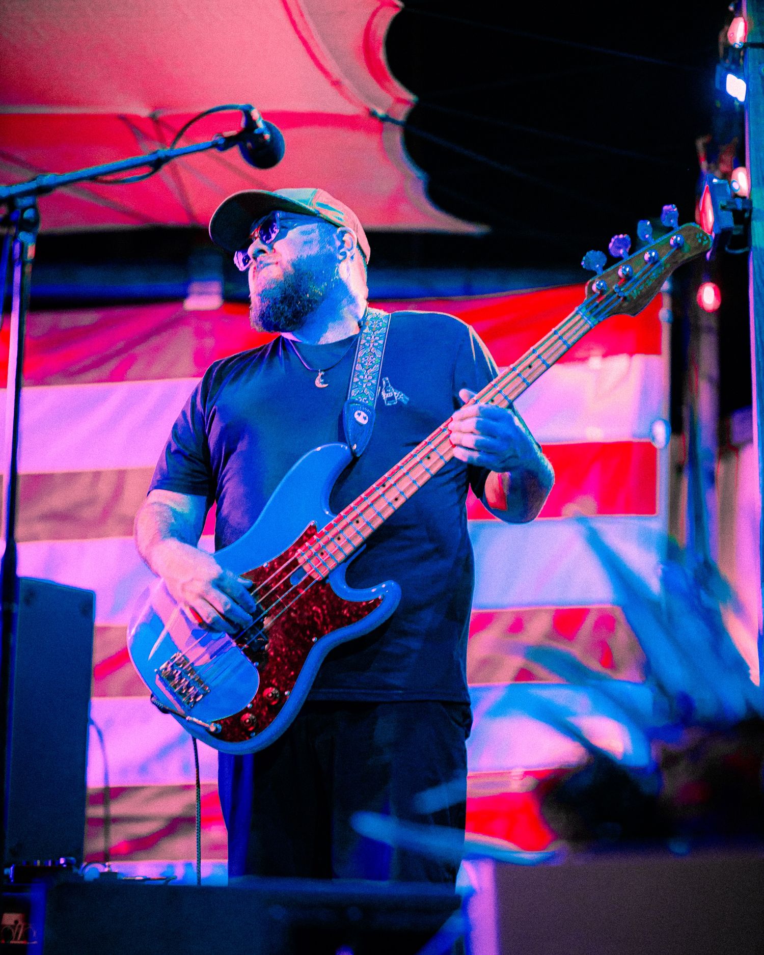 Male bassist performing on stage under colorful lights with an American flag backdrop, playing electric bass guitar at live concert.