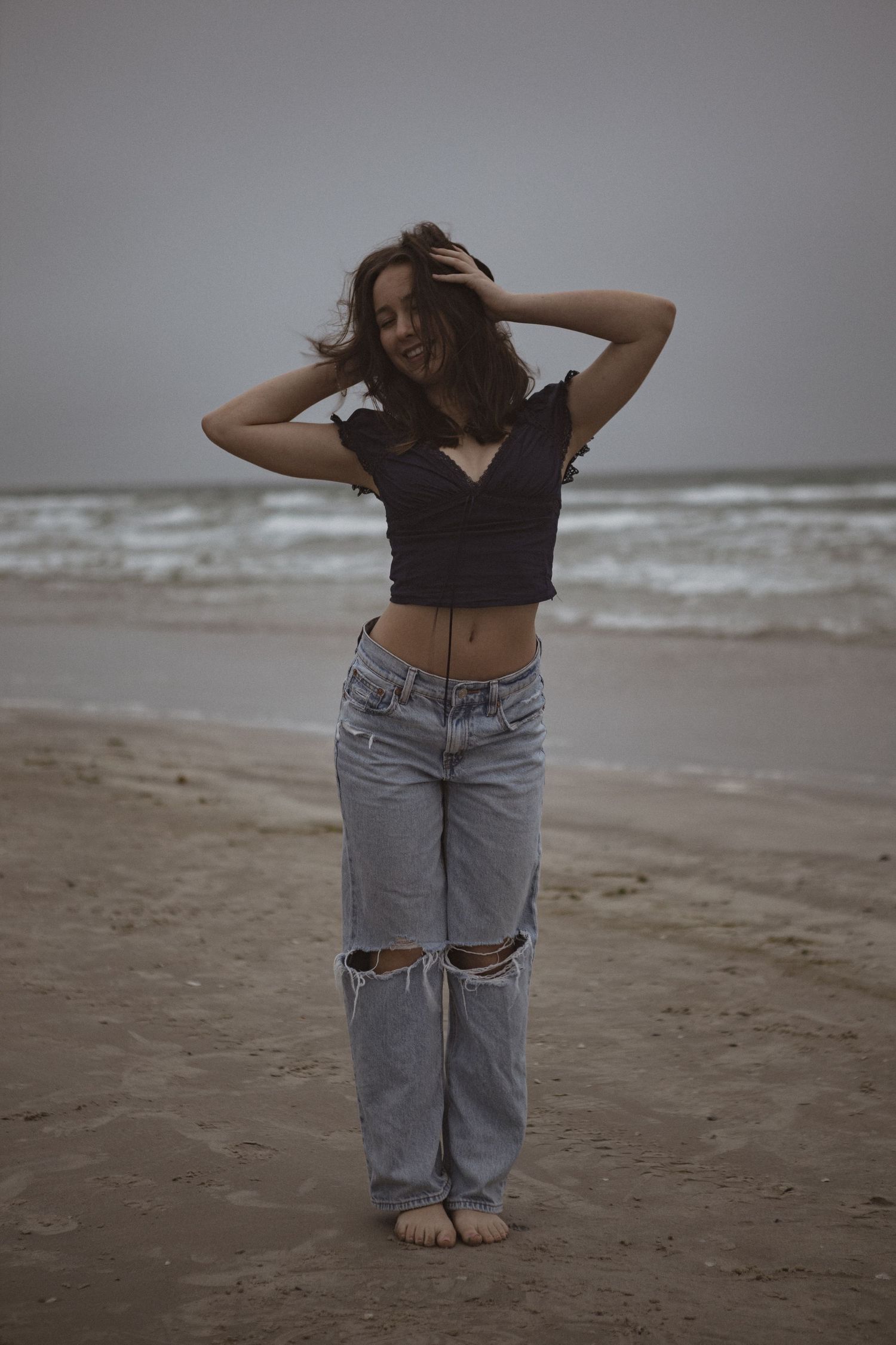 Young woman in black crop top and ripped jeans poses barefoot on a sandy beach with ocean waves behind her.