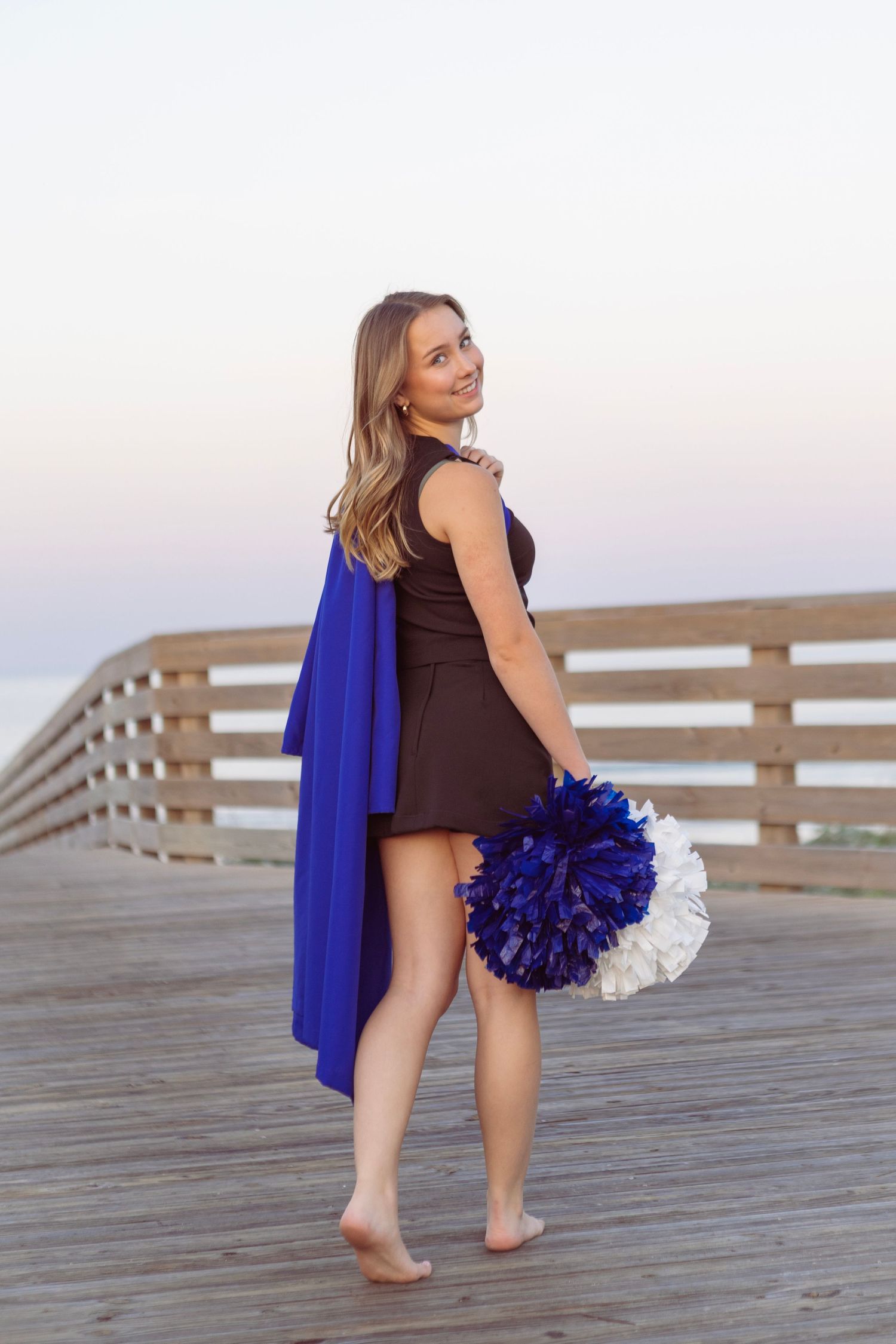 Young woman holding blue and white pom-poms on a wooden pier at dusk, wearing a brown dress with blue fabric.