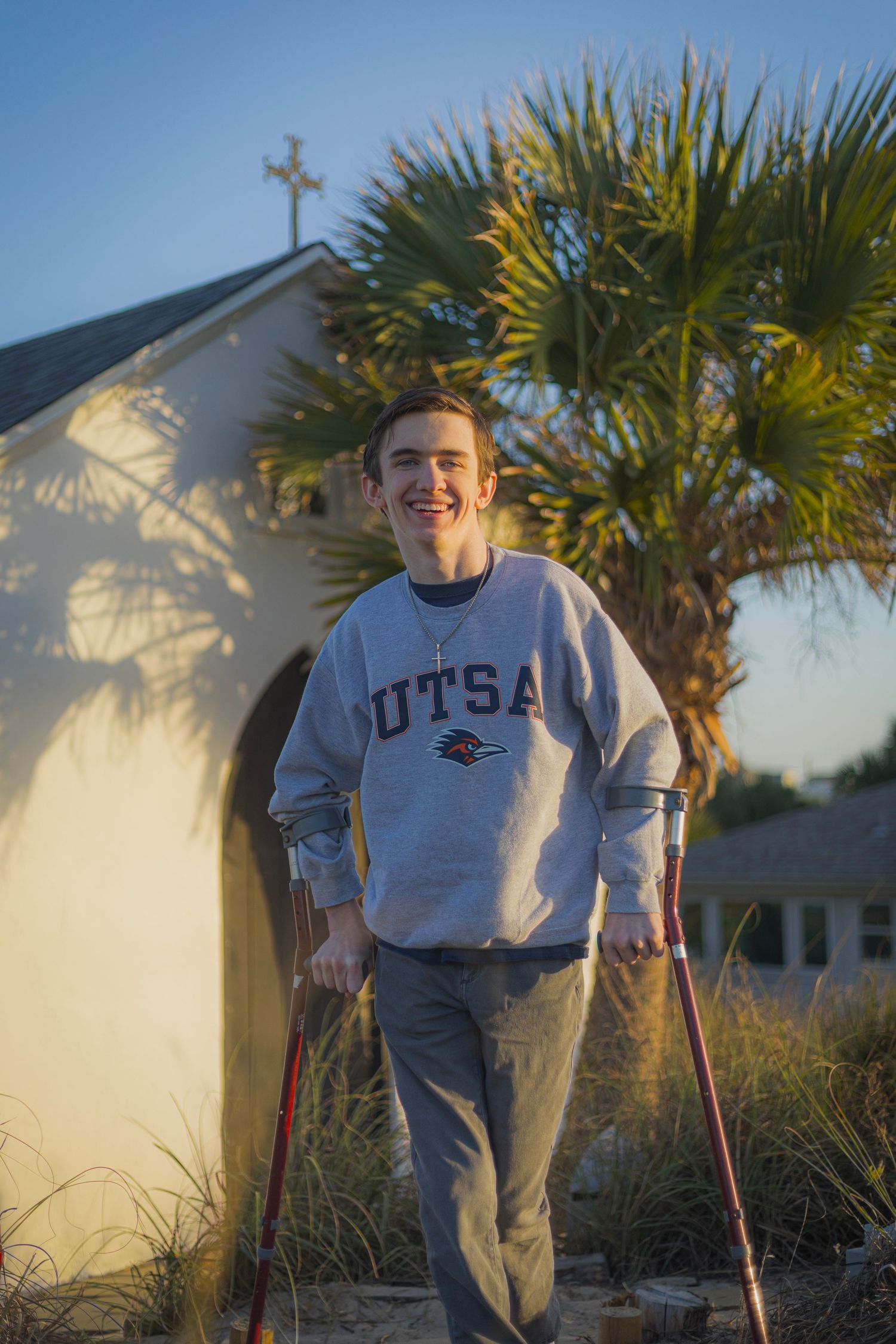 Smiling young man on crutches wearing a UTSA sweatshirt outdoors near a white building with palm trees.