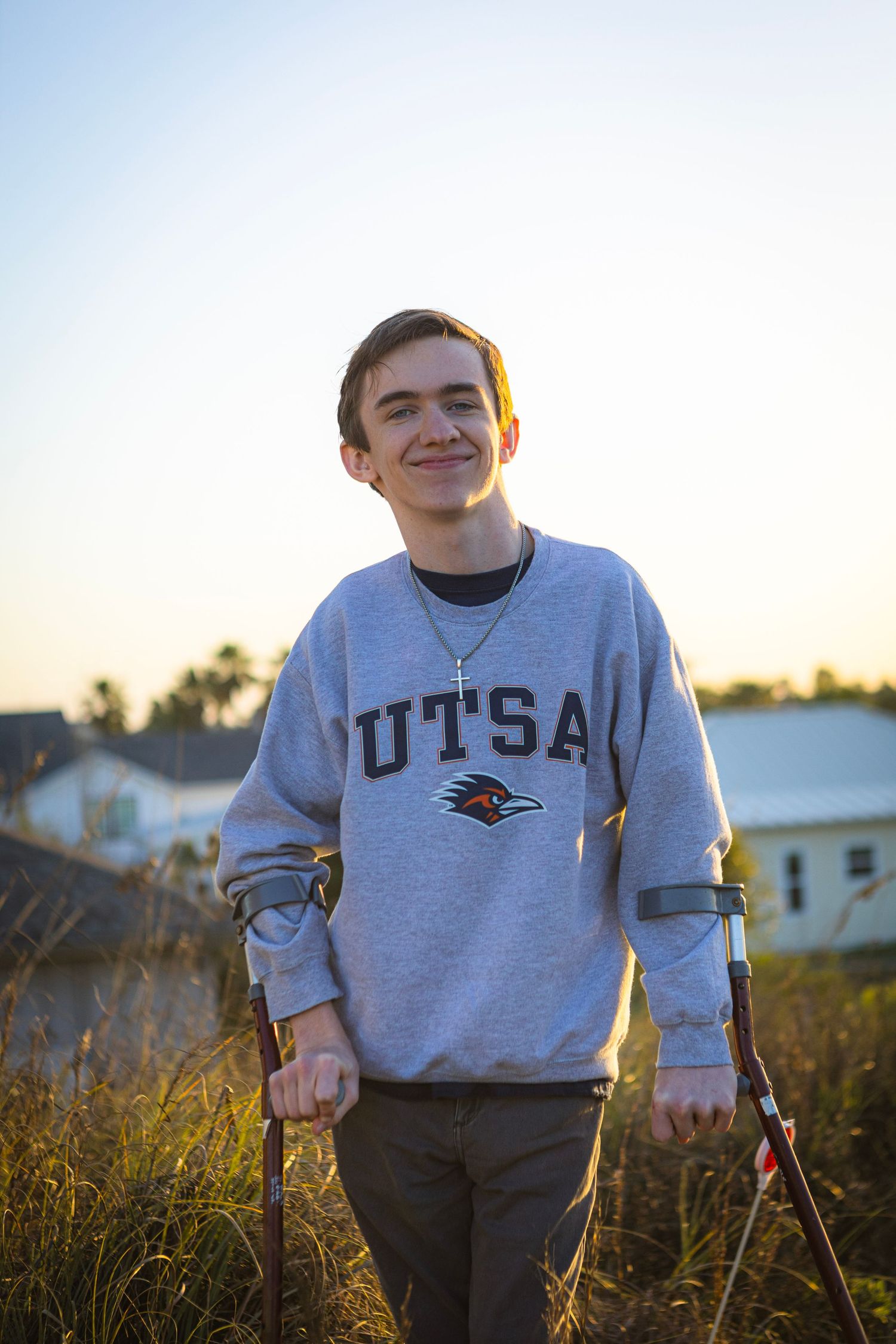 Smiling young man on forearm crutches wearing a gray UTSA sweatshirt standing in a field at golden hour.