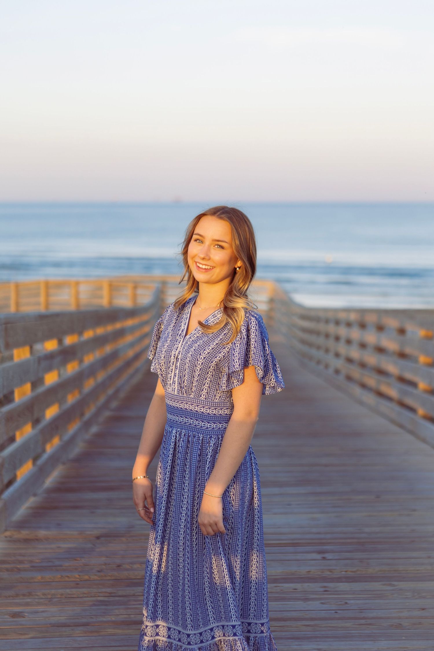 Young woman in blue floral maxi dress smiling on a wooden boardwalk pier with ocean waves in the background.