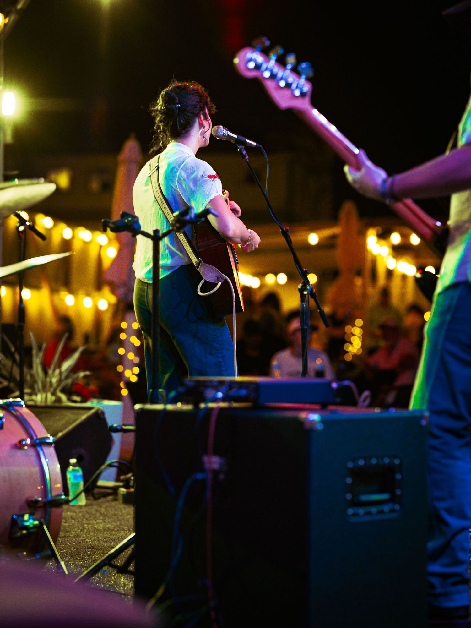 Live band performing outdoors at night with string lights, singer at microphone and guitarist on stage.