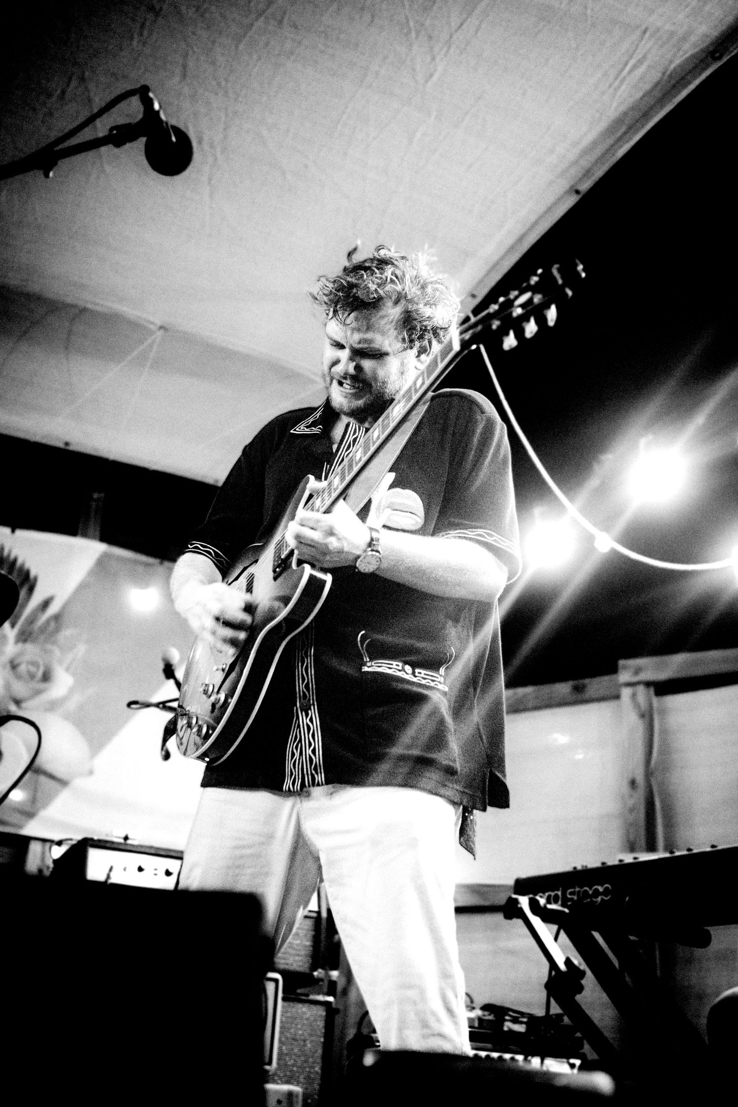 Black and white photo of a curly-haired guitarist performing on stage under bright lights with a keyboard visible nearby.