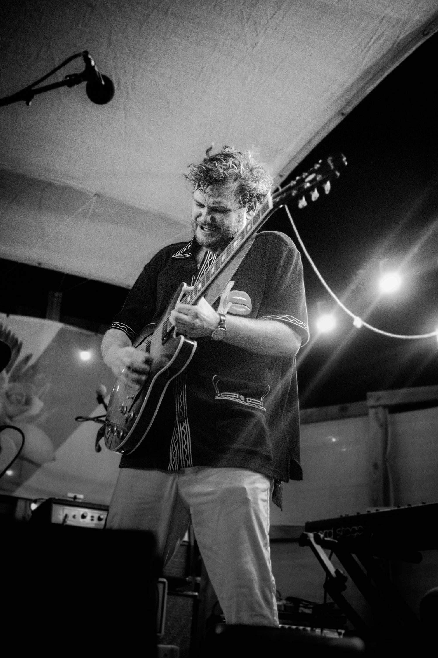 Black and white photo of a curly-haired guitarist performing on stage under string lights, playing an electric guitar.
