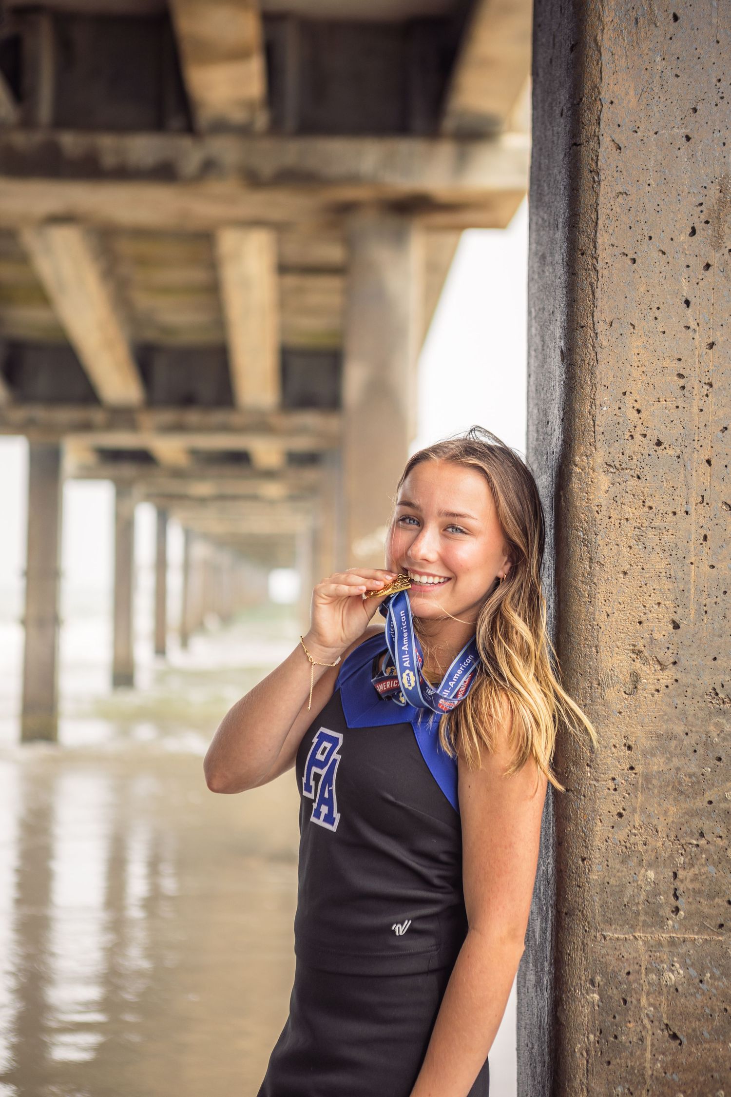 Smiling young woman in sports jersey leaning against concrete pillar under pier, wearing blue scarf and medal.