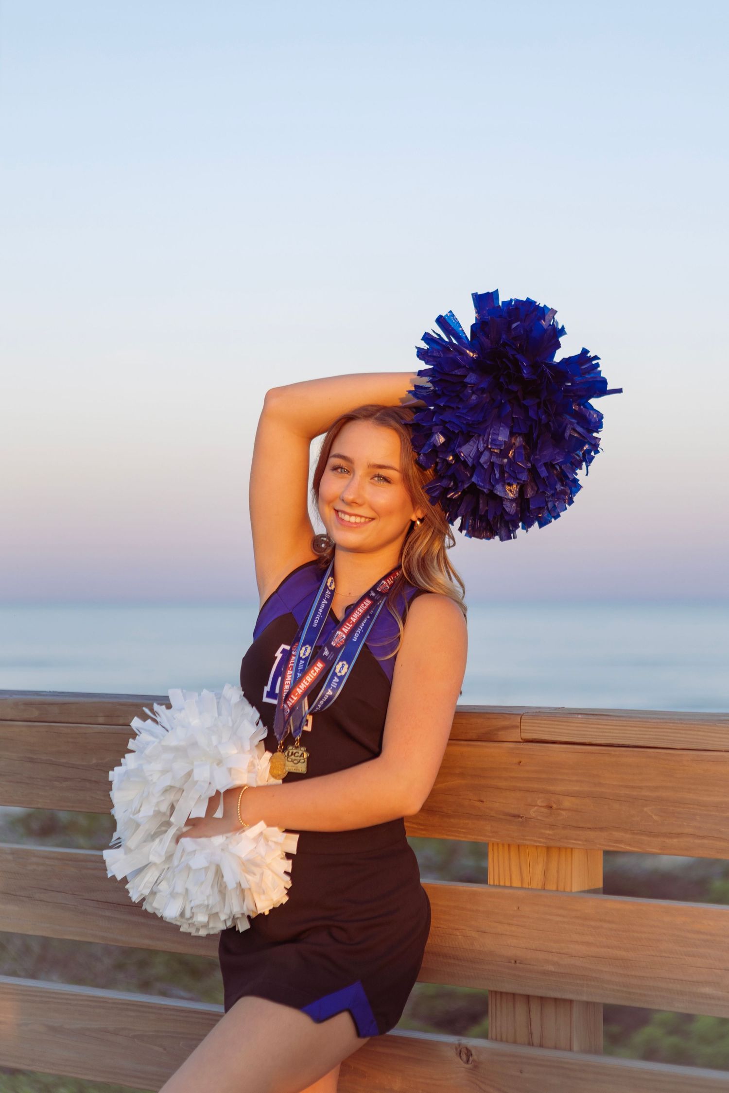 Cheerleader in uniform holding blue and white pom-poms poses on a wooden boardwalk at sunset near the ocean.