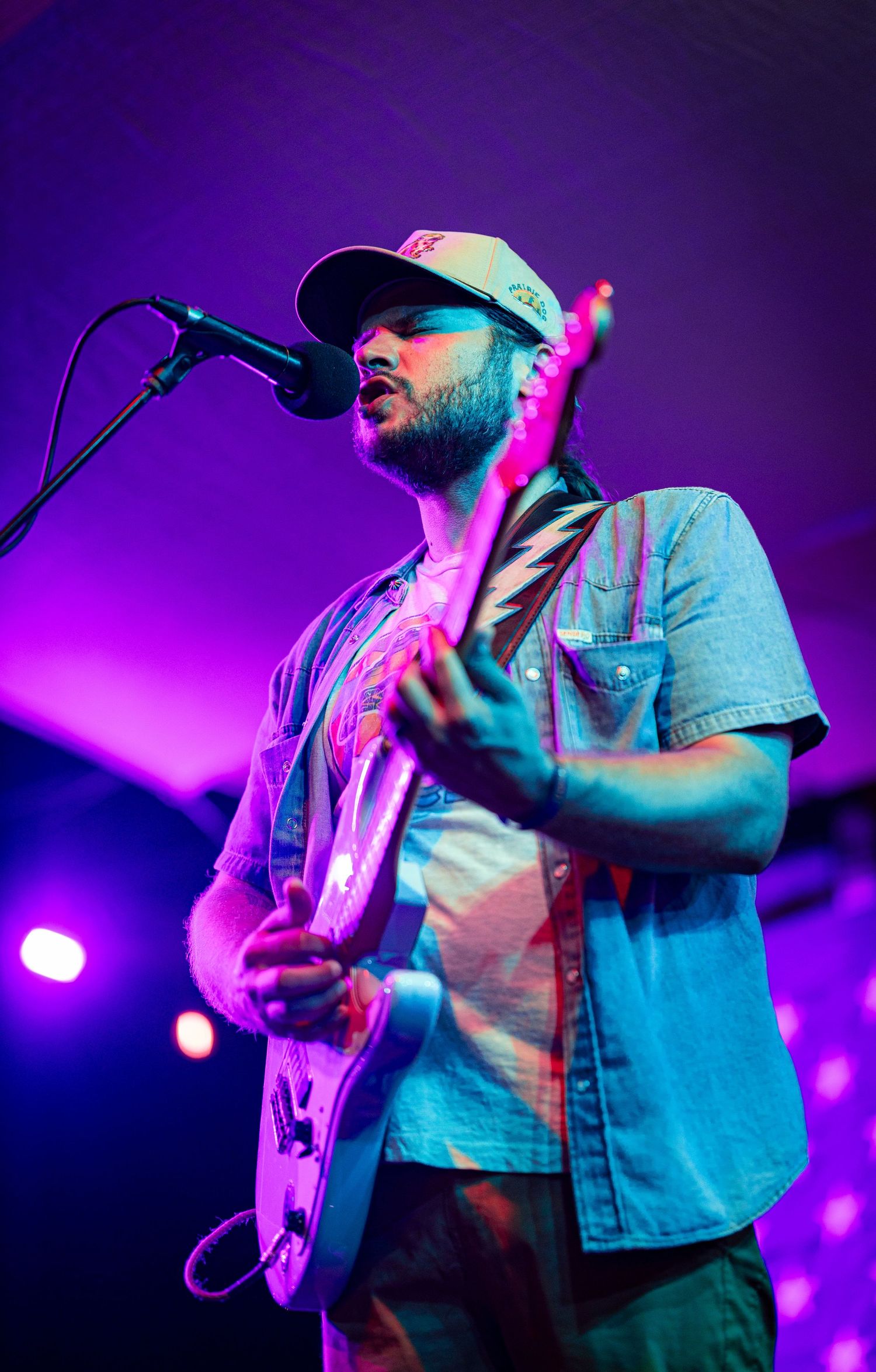 Bearded male guitarist performing on stage under purple lights, wearing a cap and denim shirt while singing into a microphone.