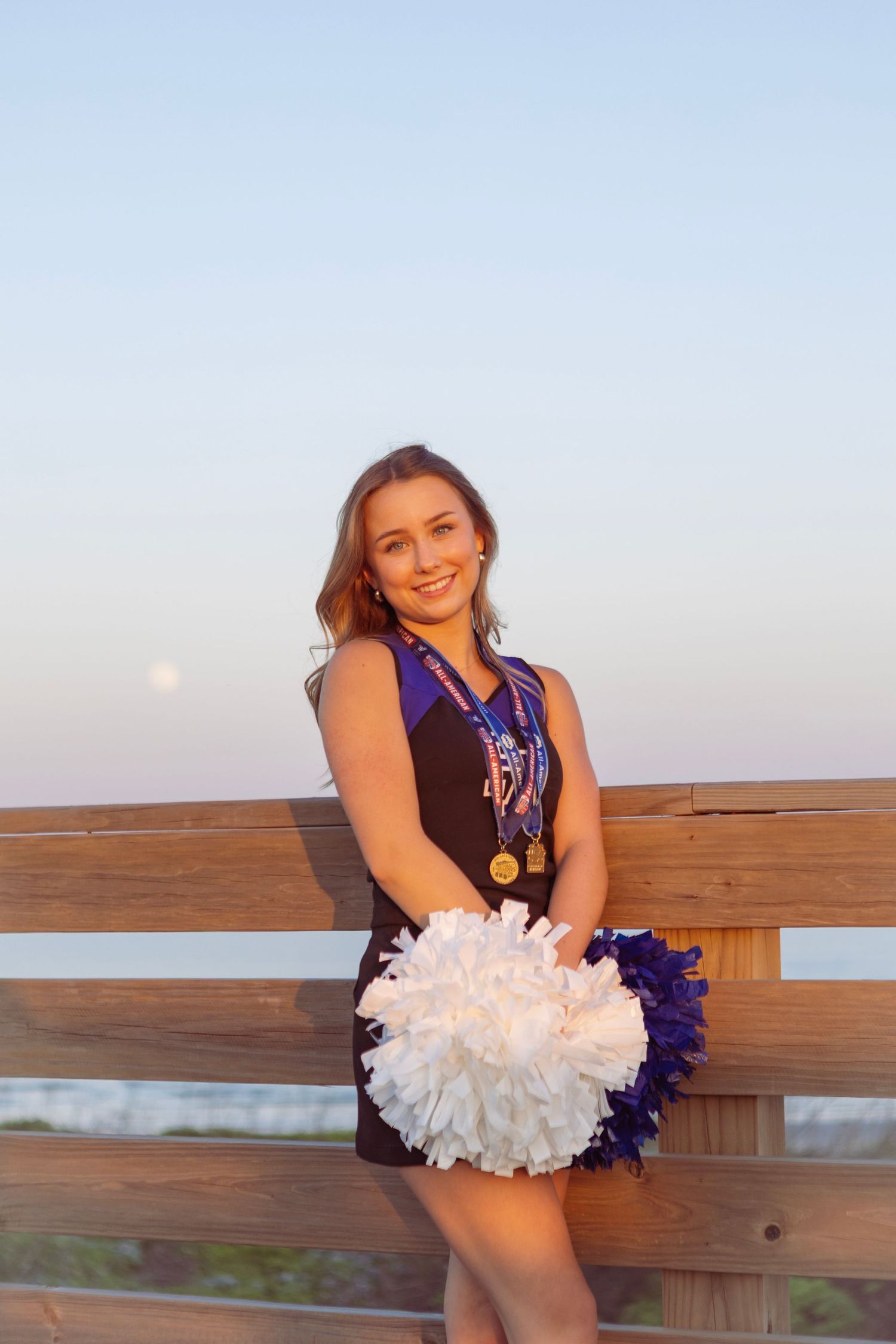 Young cheerleader in uniform holding white and purple pom-poms, wearing gold medals, posing on wooden bridge at sunset.