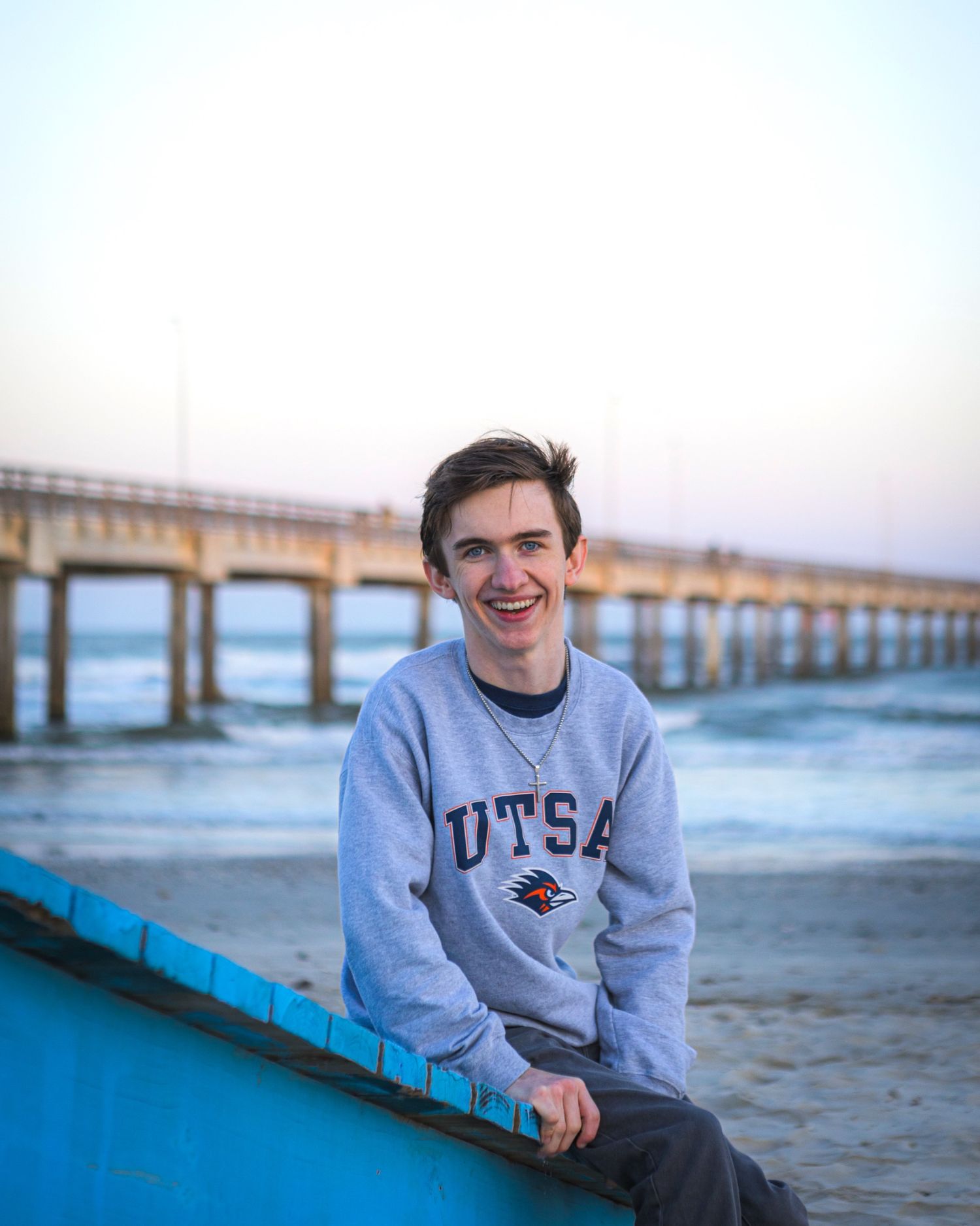Smiling young man in UTSA sweatshirt sitting on blue boat at beach with pier in background at sunset.
