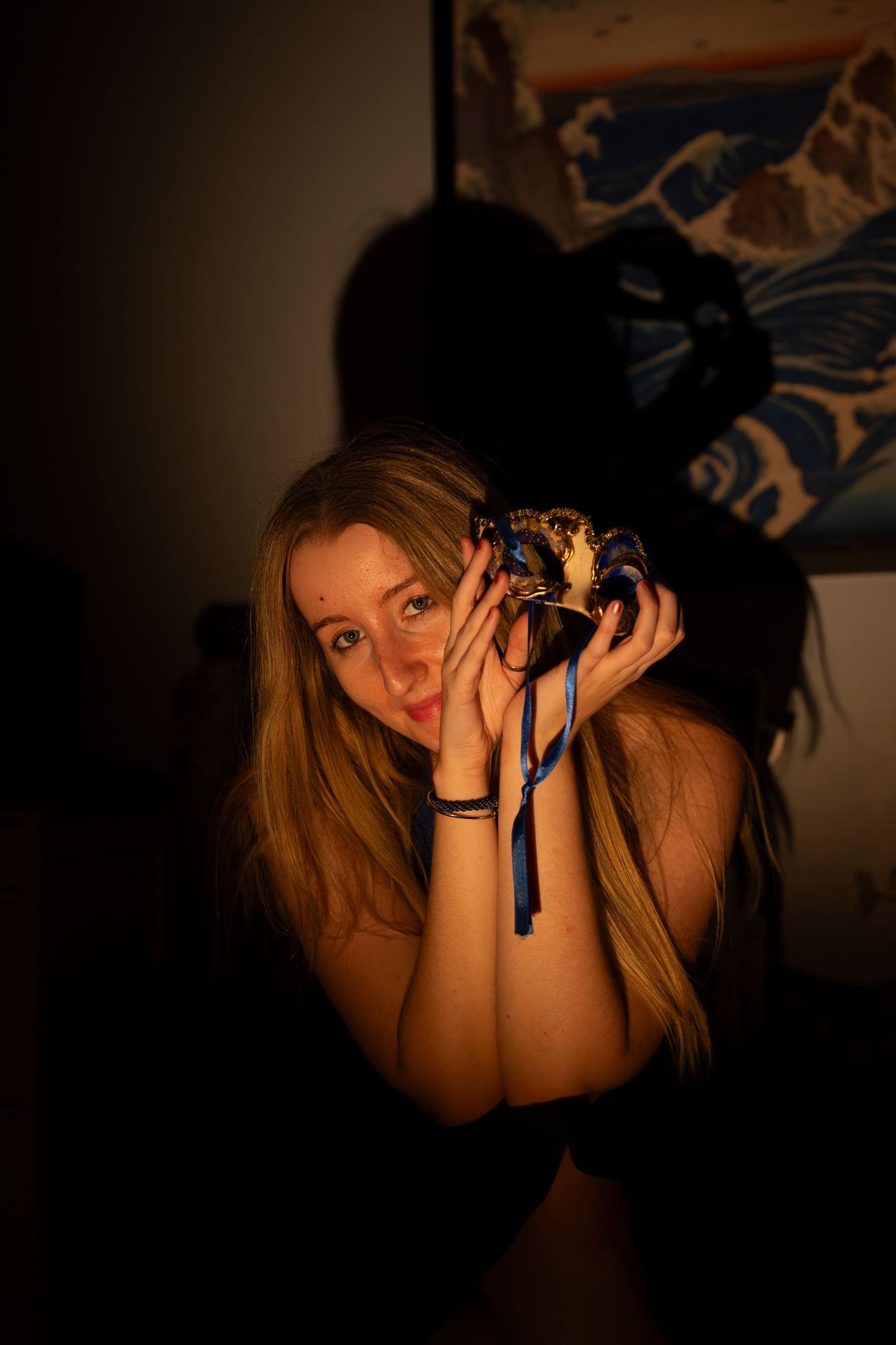 Young woman with long blonde hair holds a decorative Venetian masquerade mask in dim, warm lighting.
