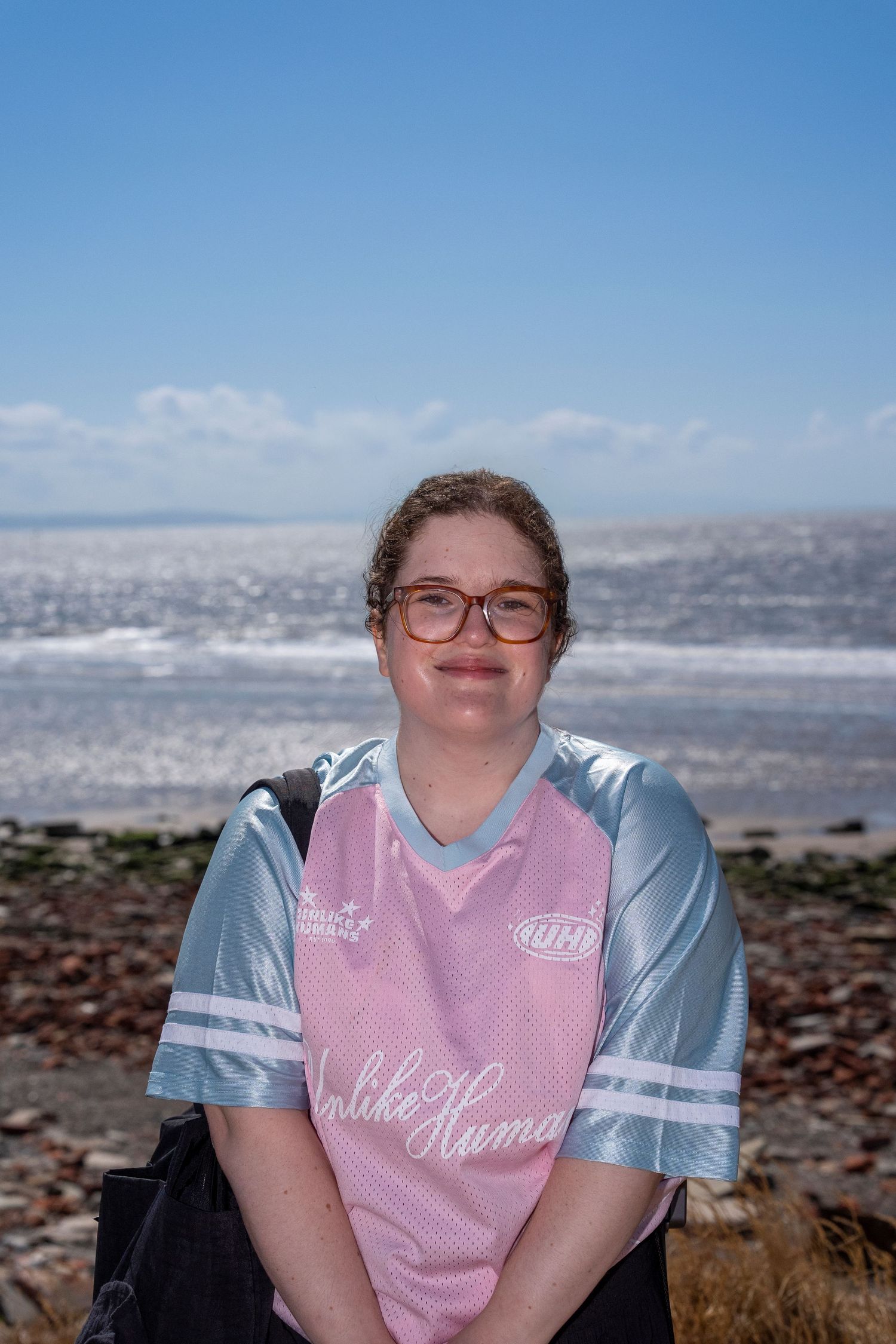 Young woman with glasses wearing a pink and silver jersey smiles at camera with ocean beach backdrop on sunny day.