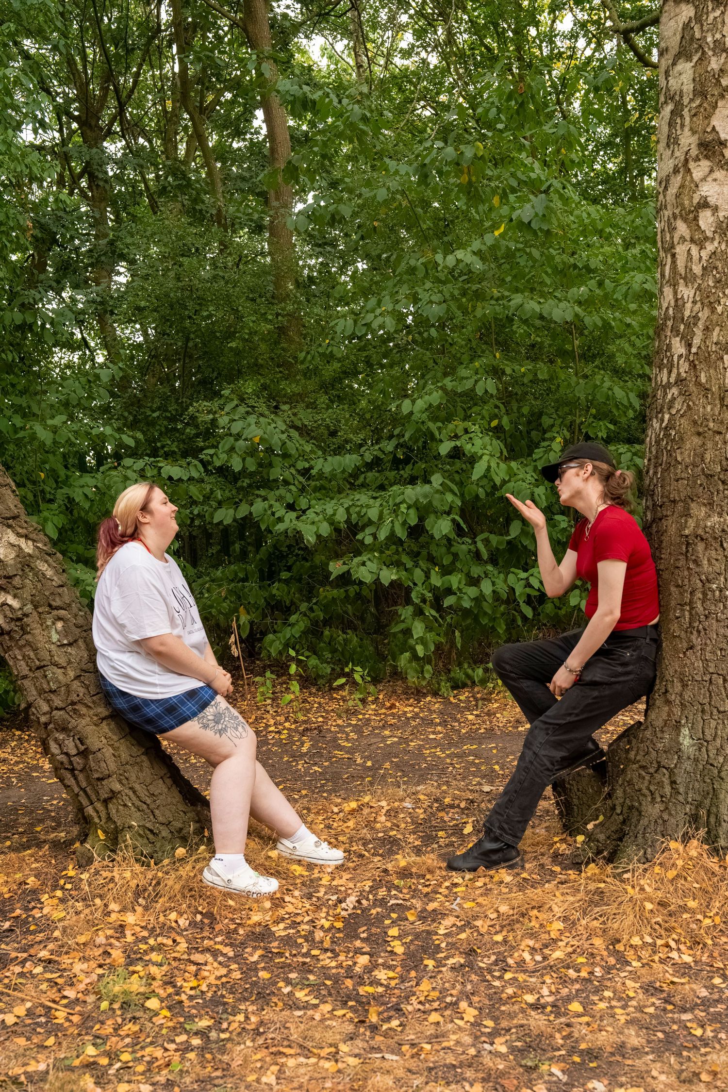 Two women having a conversation while leaning against trees in an autumn forest park setting.