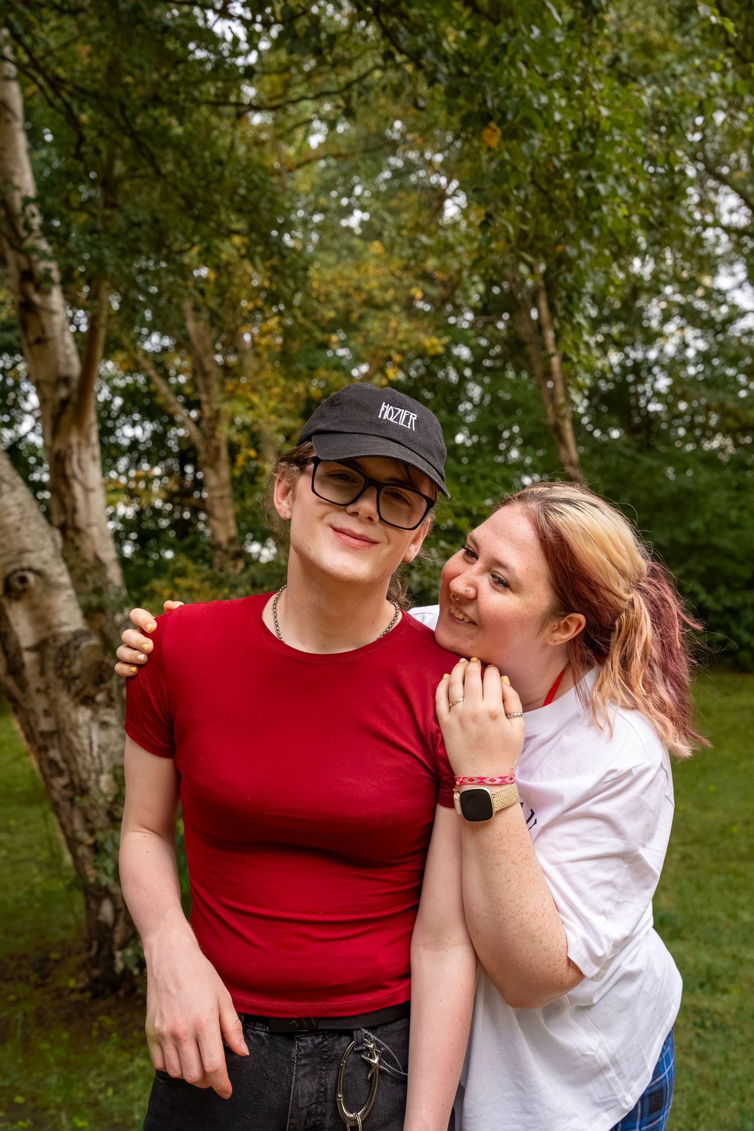 Two young women smiling and embracing outdoors in a park, one in a red shirt and cap, the other in a white tee.