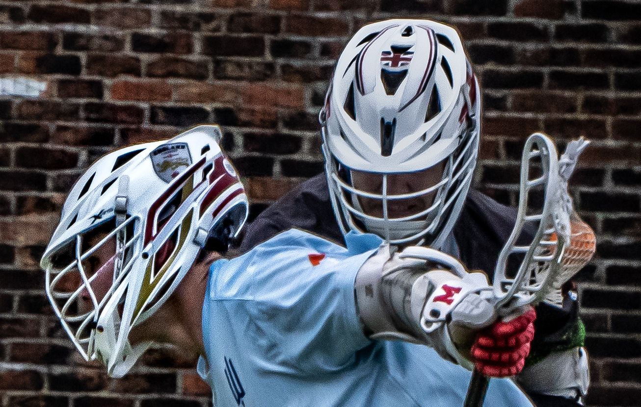 Two lacrosse players in white helmets facing off closely against a brick wall background.