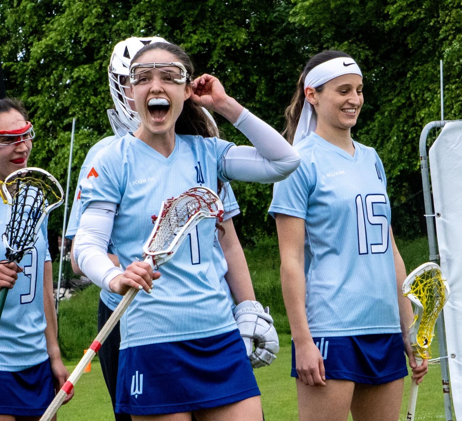 Female lacrosse players in light blue uniforms walking onto the field, one wearing goggles holding a stick.