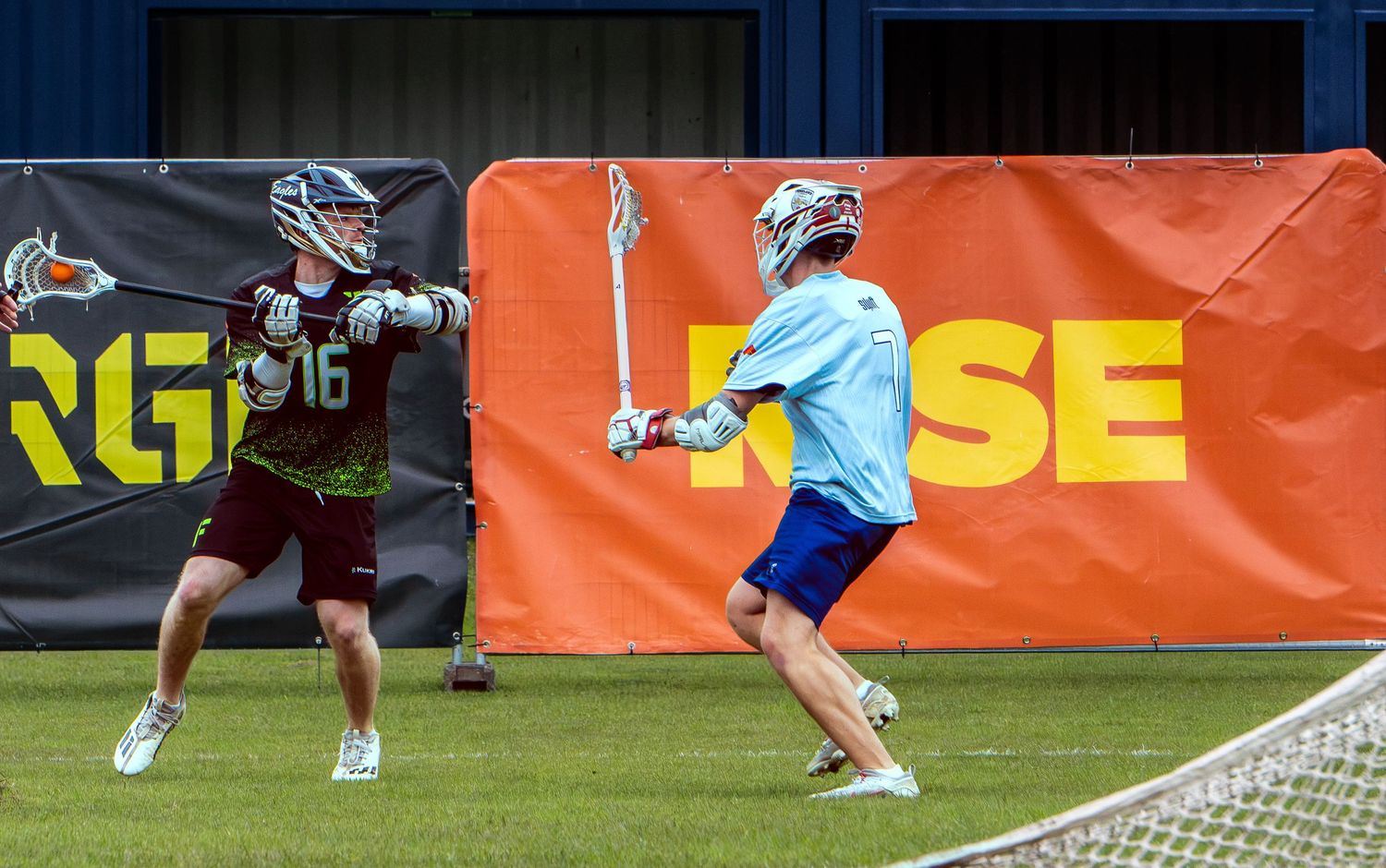 Two lacrosse players competing during a practice match in front of an orange advertising banner.