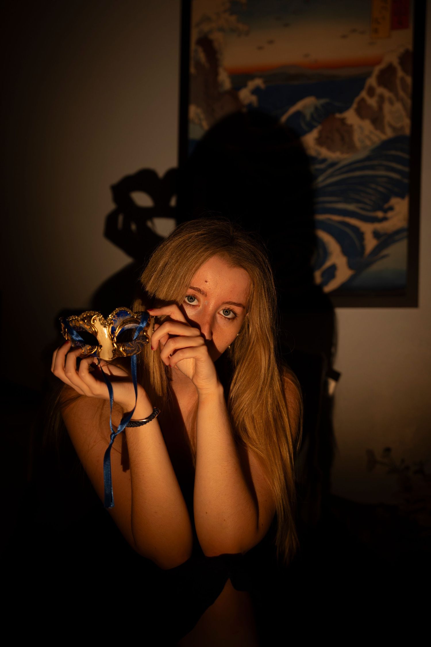 Blonde woman holding a decorative masquerade mask in dim warm lighting with a Japanese art print on the wall.