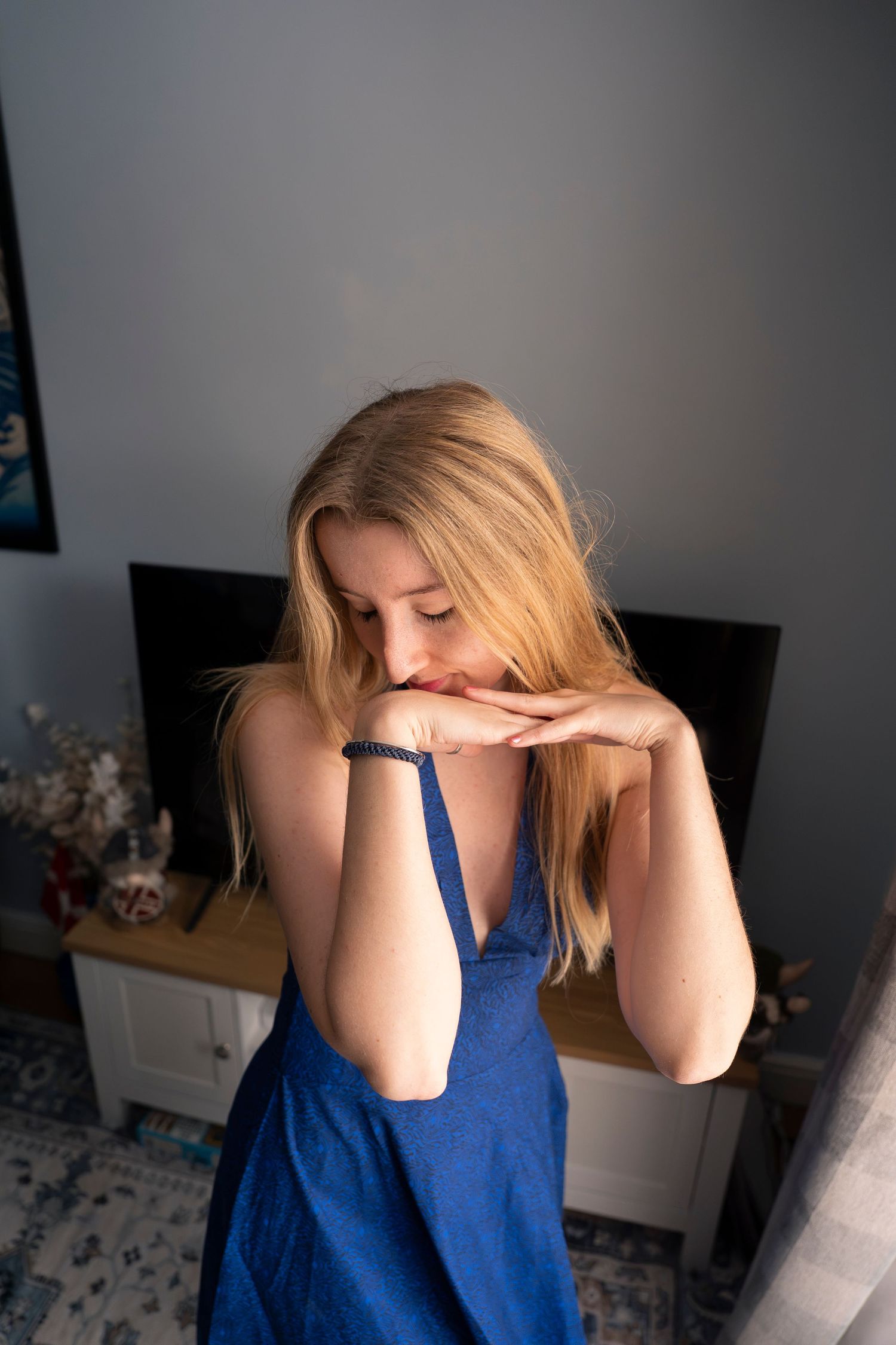 Young blonde woman in blue dress resting chin on hands, looking down thoughtfully in a softly lit room.