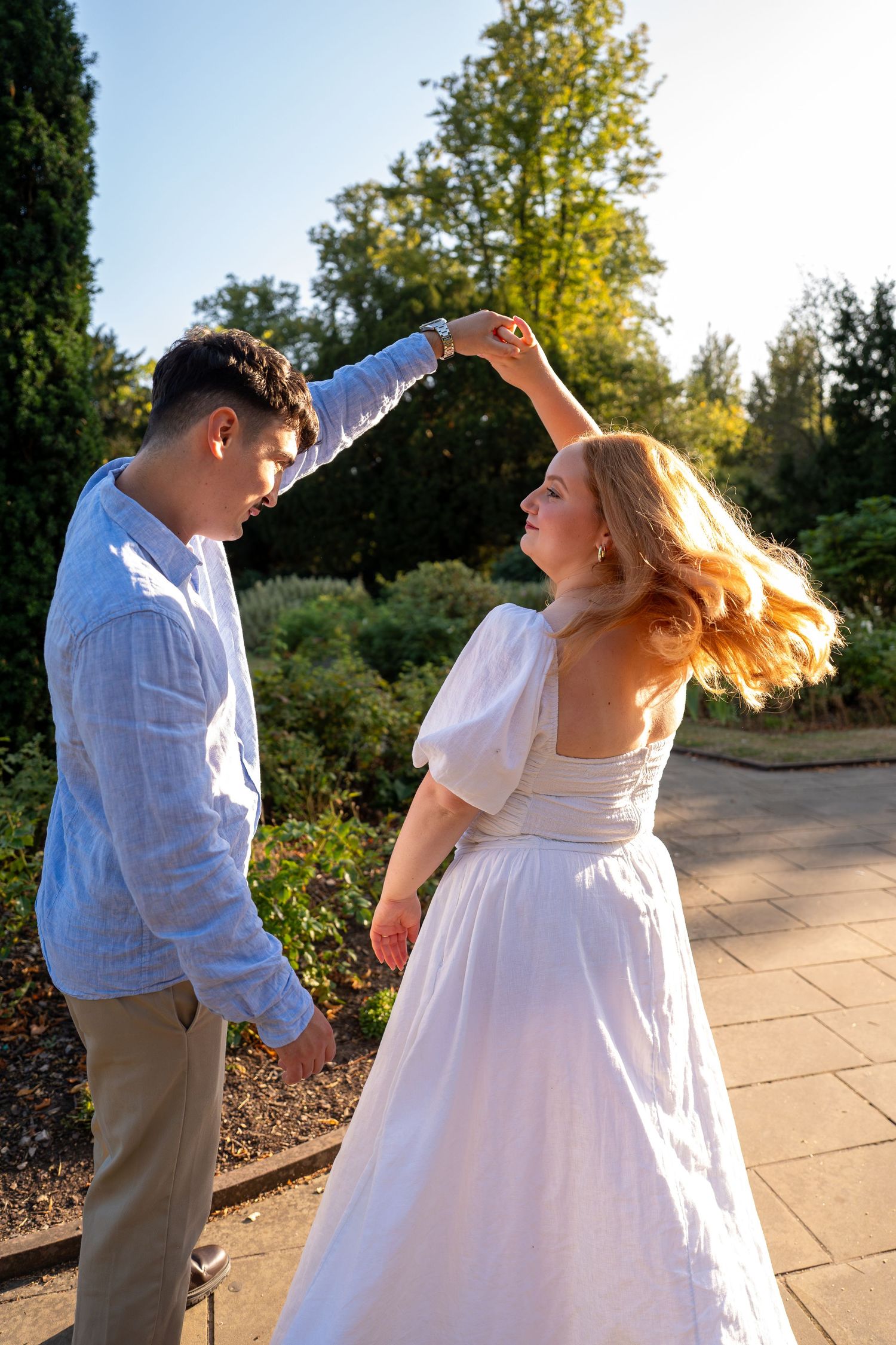 A couple dancing outdoors in a sunny garden, woman in white dress being twirled by man in blue shirt.