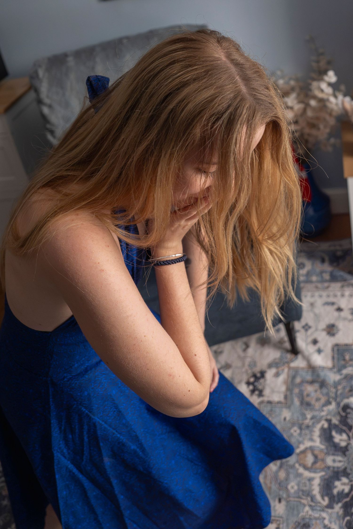 Young blonde woman in blue dress sitting on floor with head down, appearing sad or distressed indoors.