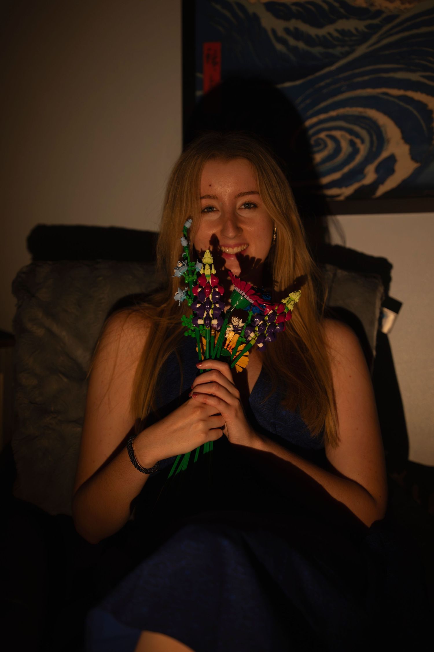 Smiling blonde woman holds colorful wildflower bouquet in cozy amber-lit room with wave art.