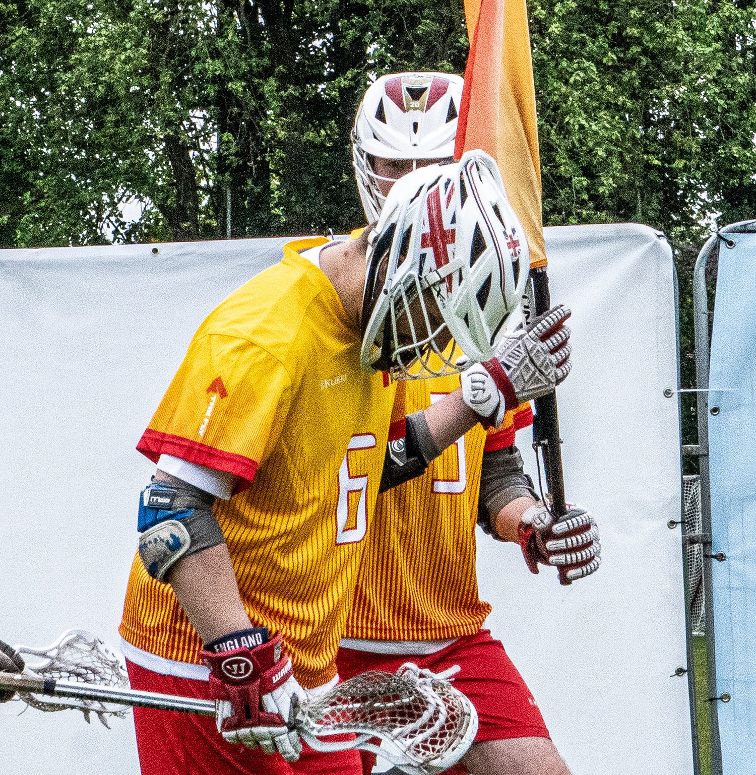 Male Lacrosse player in a yellow and red uniform wearing a white helmet holding a stick.