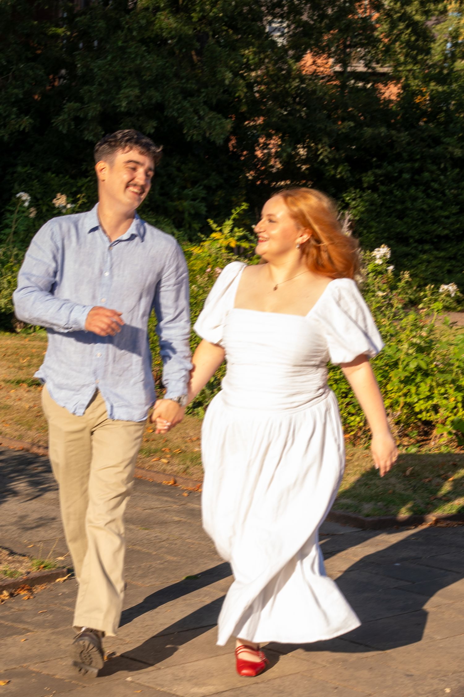 A smiling couple holds hands while walking in a sunny park, woman in white dress and man in blue shirt.